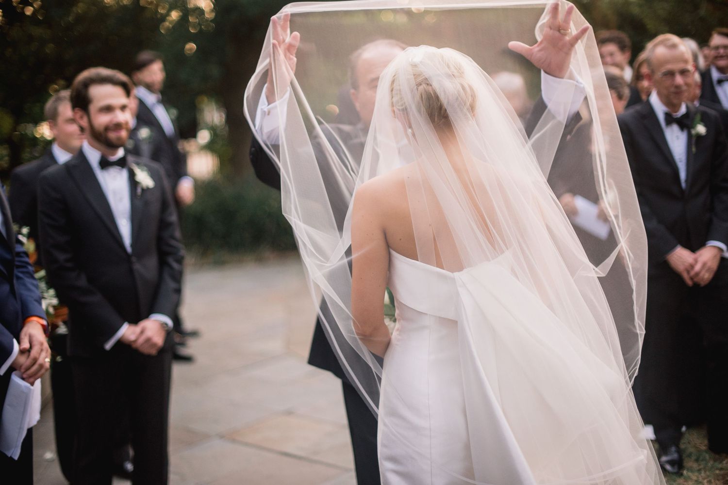 A bride's veil flows gracefully in the breeze during an outdoor wedding ceremony with guests looking on.