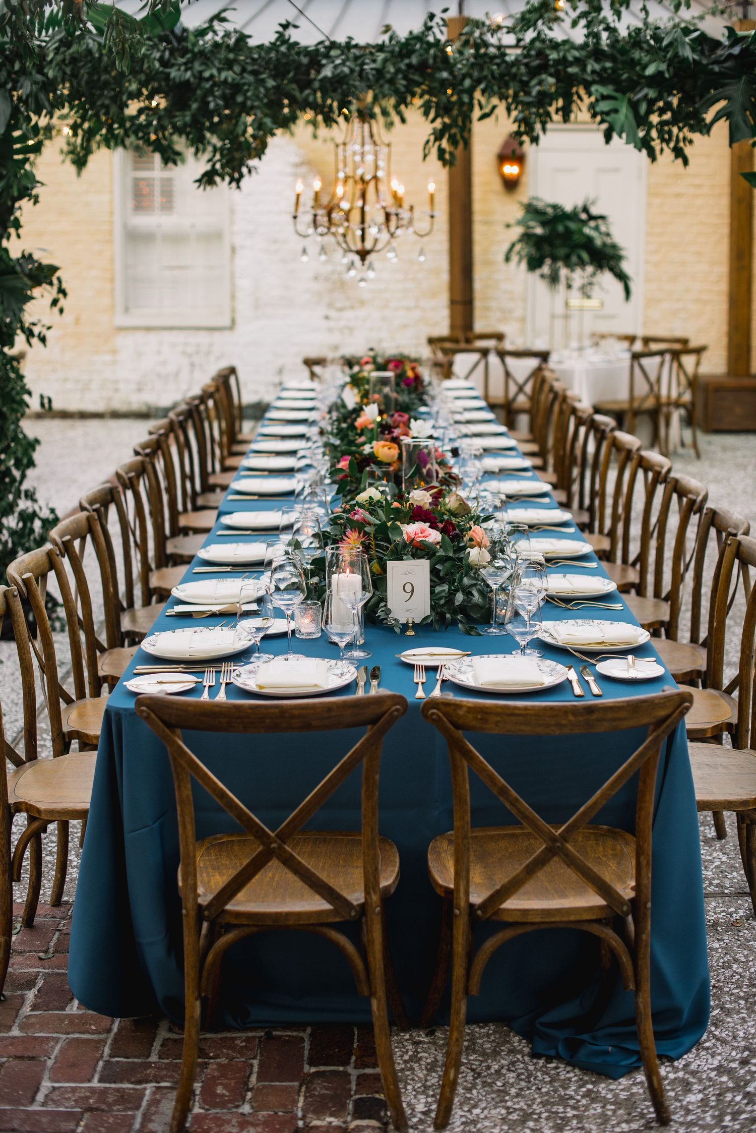 Elegant wedding reception table setup with navy blue linens, rustic wooden chairs, and greenery garland centerpieces.