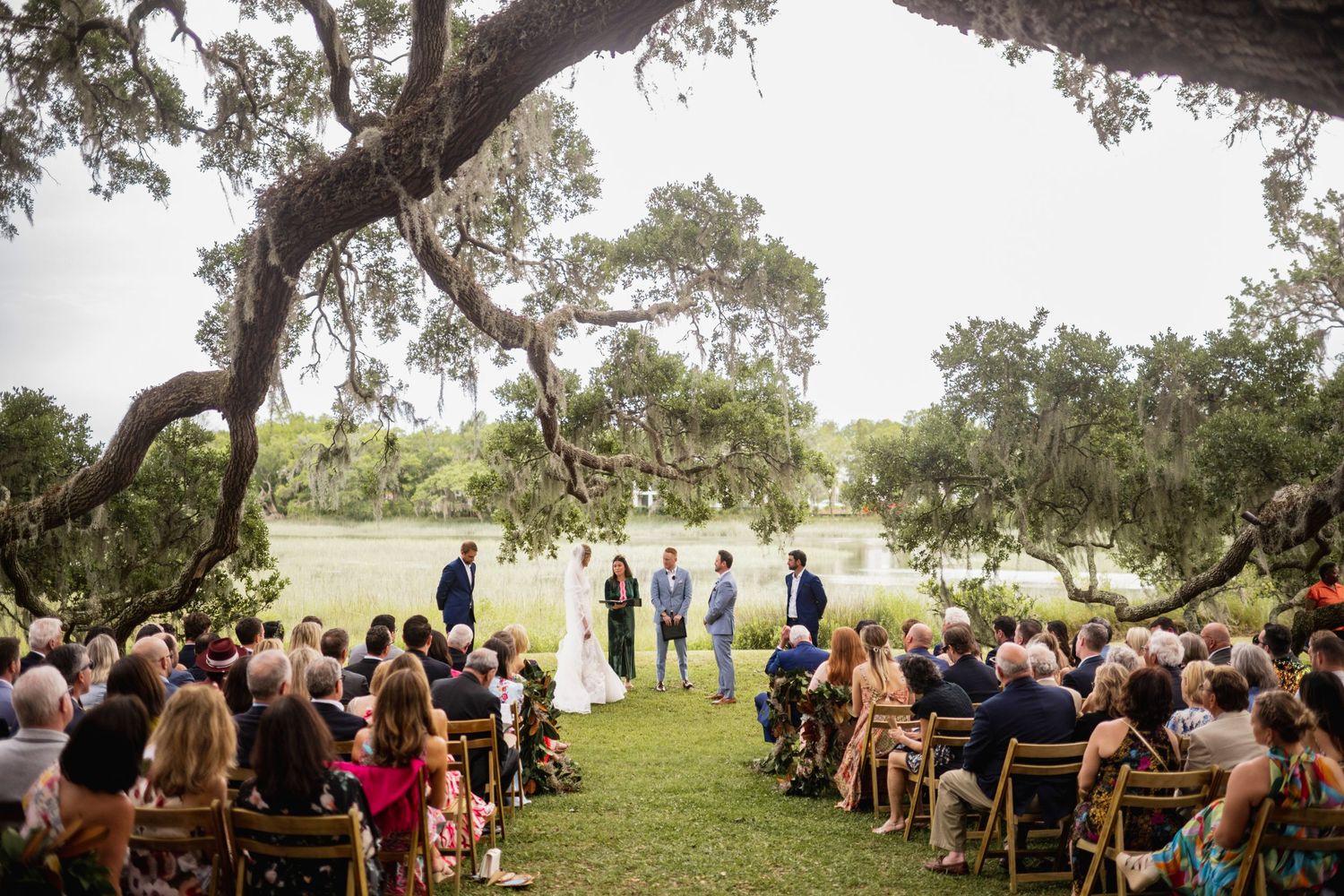 Outdoor wedding ceremony under large oak trees with guests seated in wooden chairs on a grassy lawn.