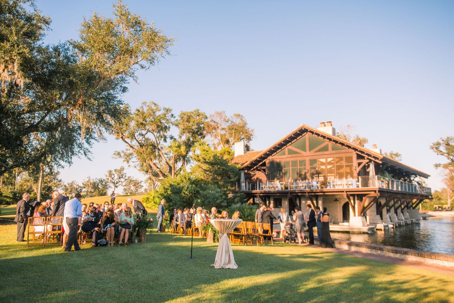 A panoramic view of a rustic wooden lodge venue during a sunset wedding ceremony with guests gathered on the lawn.