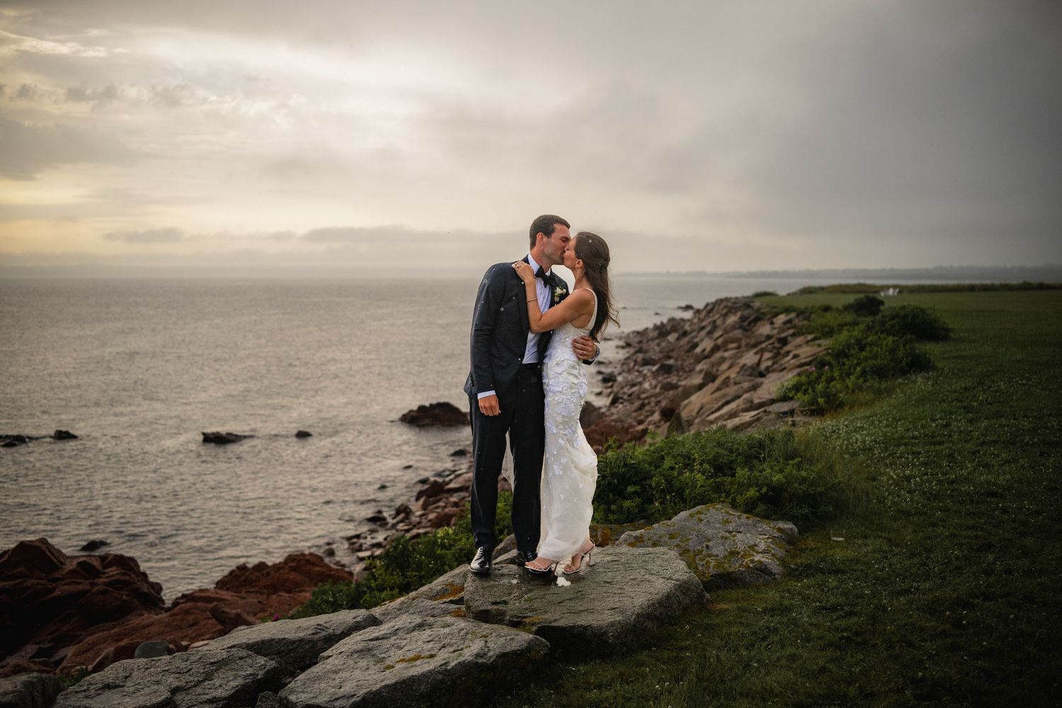 A couple shares a romantic moment on a rocky coastal cliff during sunset while wearing formal wedding attire.