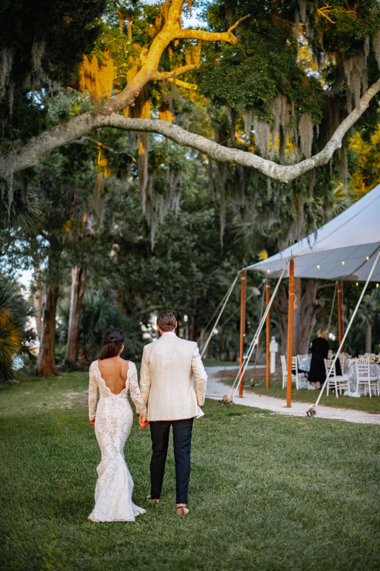 Couple walking away together on grassy lawn with white tent and Spanish moss draped trees at outdoor wedding venue.