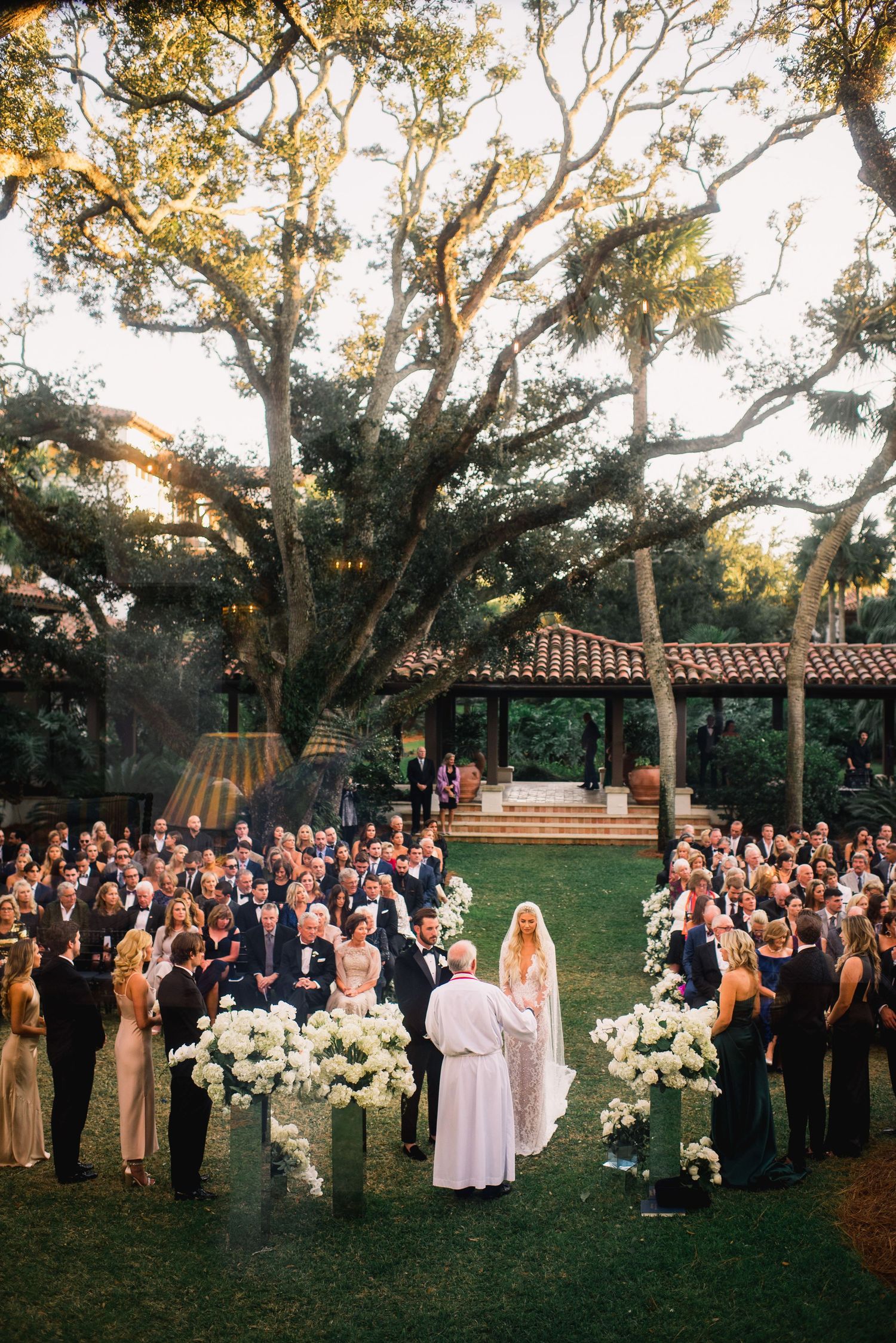 Wedding guests gather outdoors under large oak trees at sunset during an elegant garden ceremony with white floral arrangements.