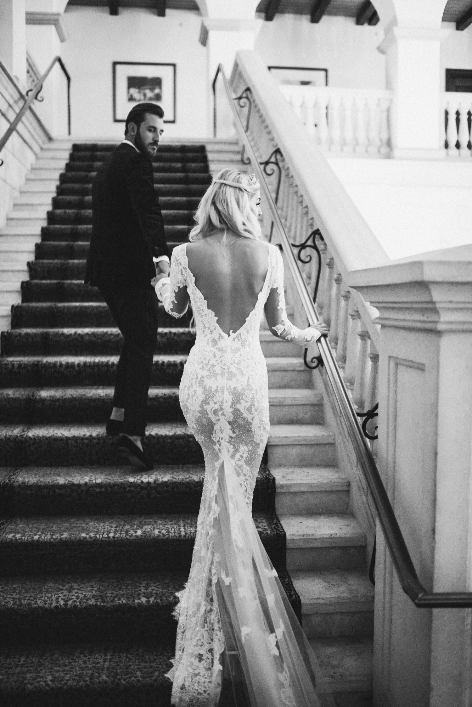 A bride in an elegant lace wedding gown walks up a grand white staircase in a classic black and white photograph.