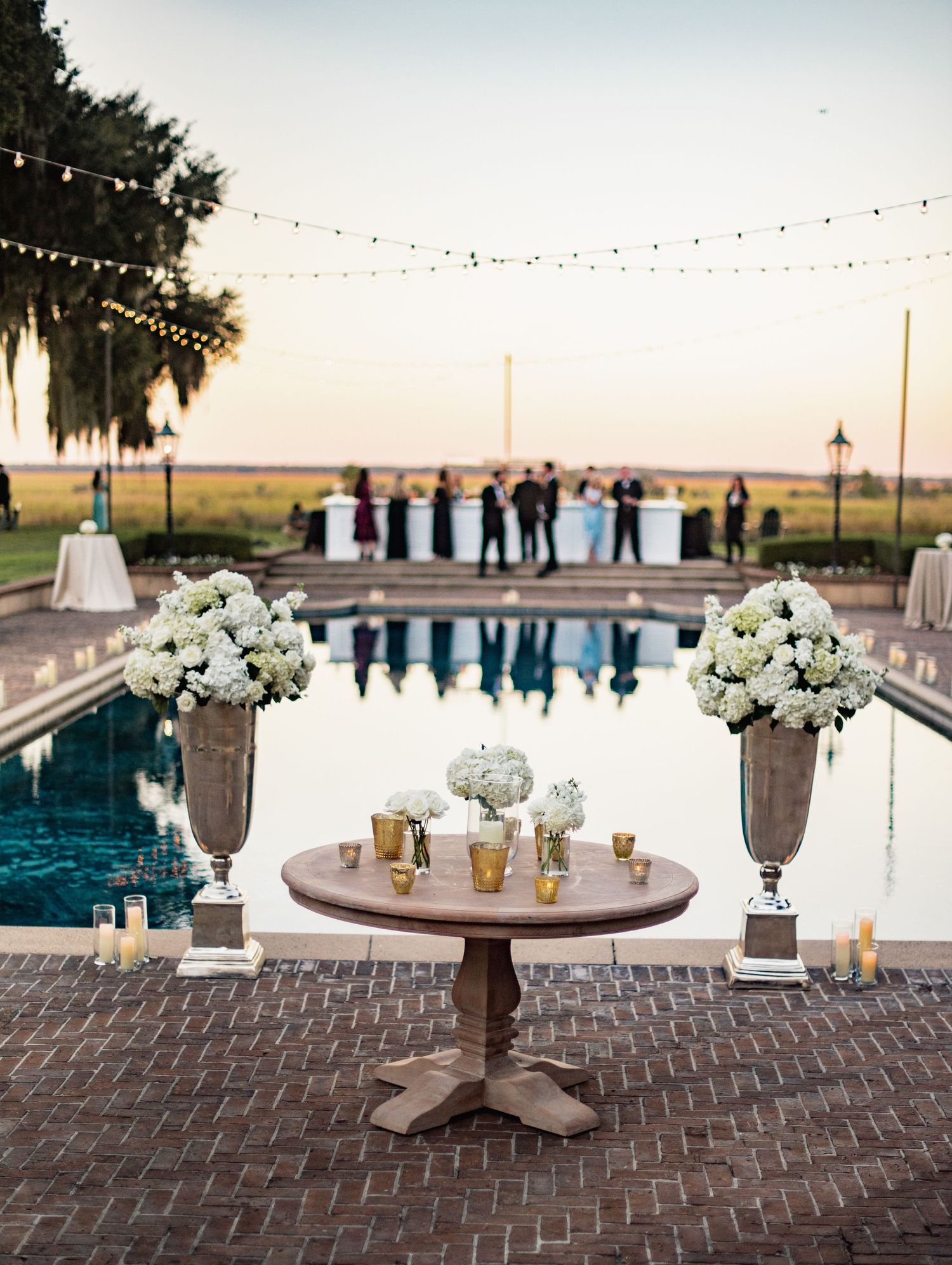 Elegant outdoor wedding reception setup with string lights, pool views, and white floral centerpieces on brick patio at sunset.