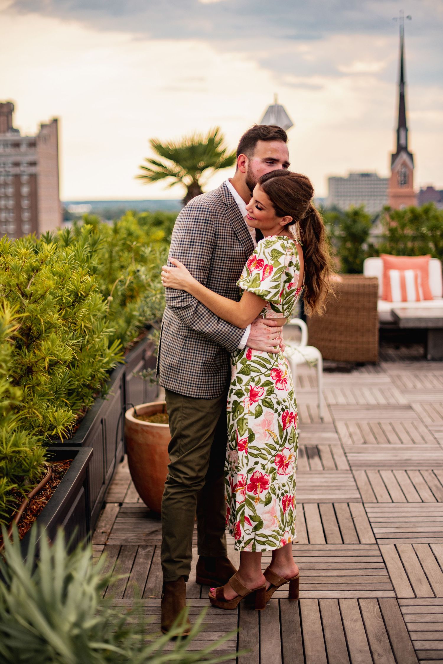 A couple embraces romantically on an urban rooftop deck with greenery and city architecture in the background.