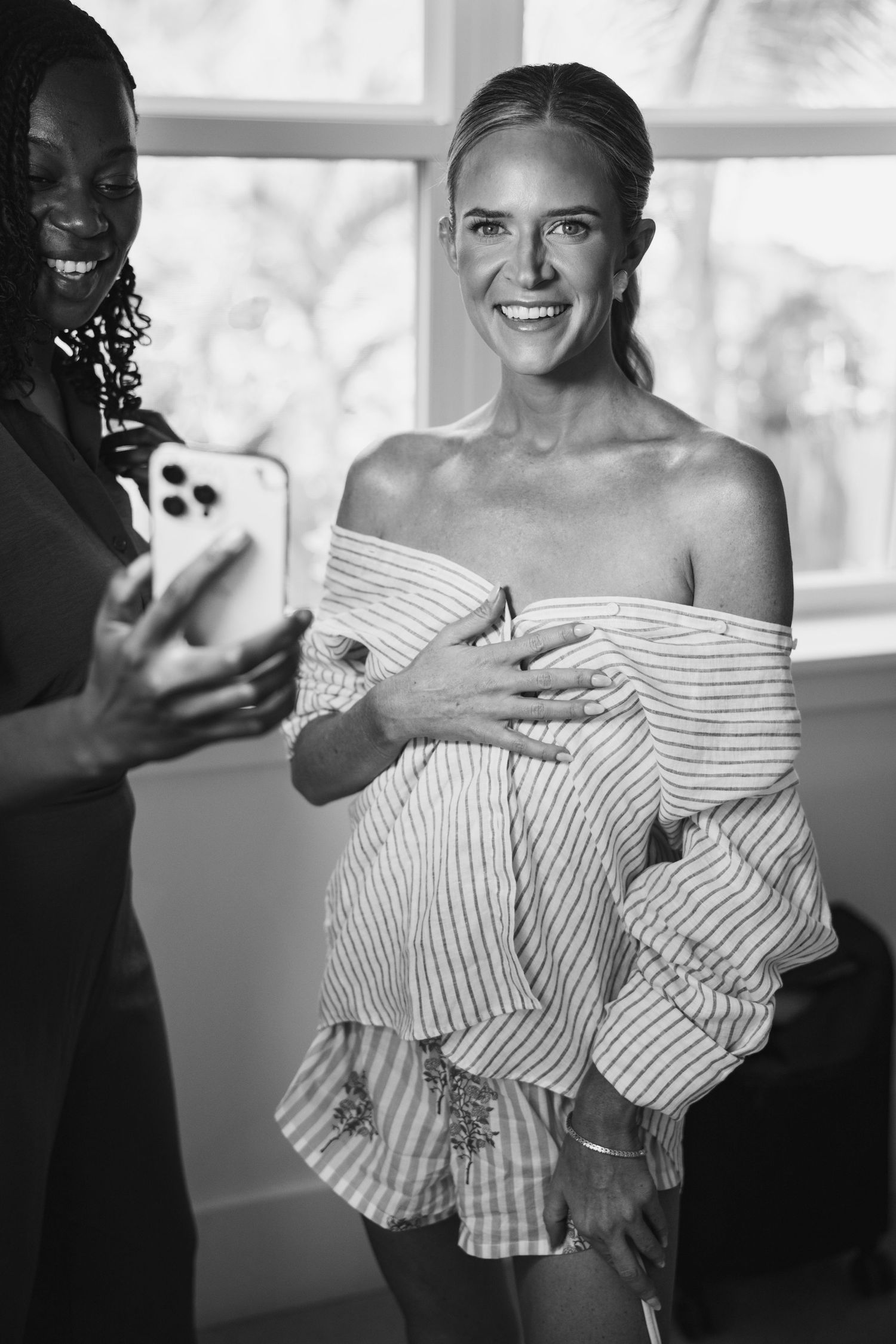 Two people share a candid black and white moment while looking at a phone, one wearing an off-shoulder striped top.