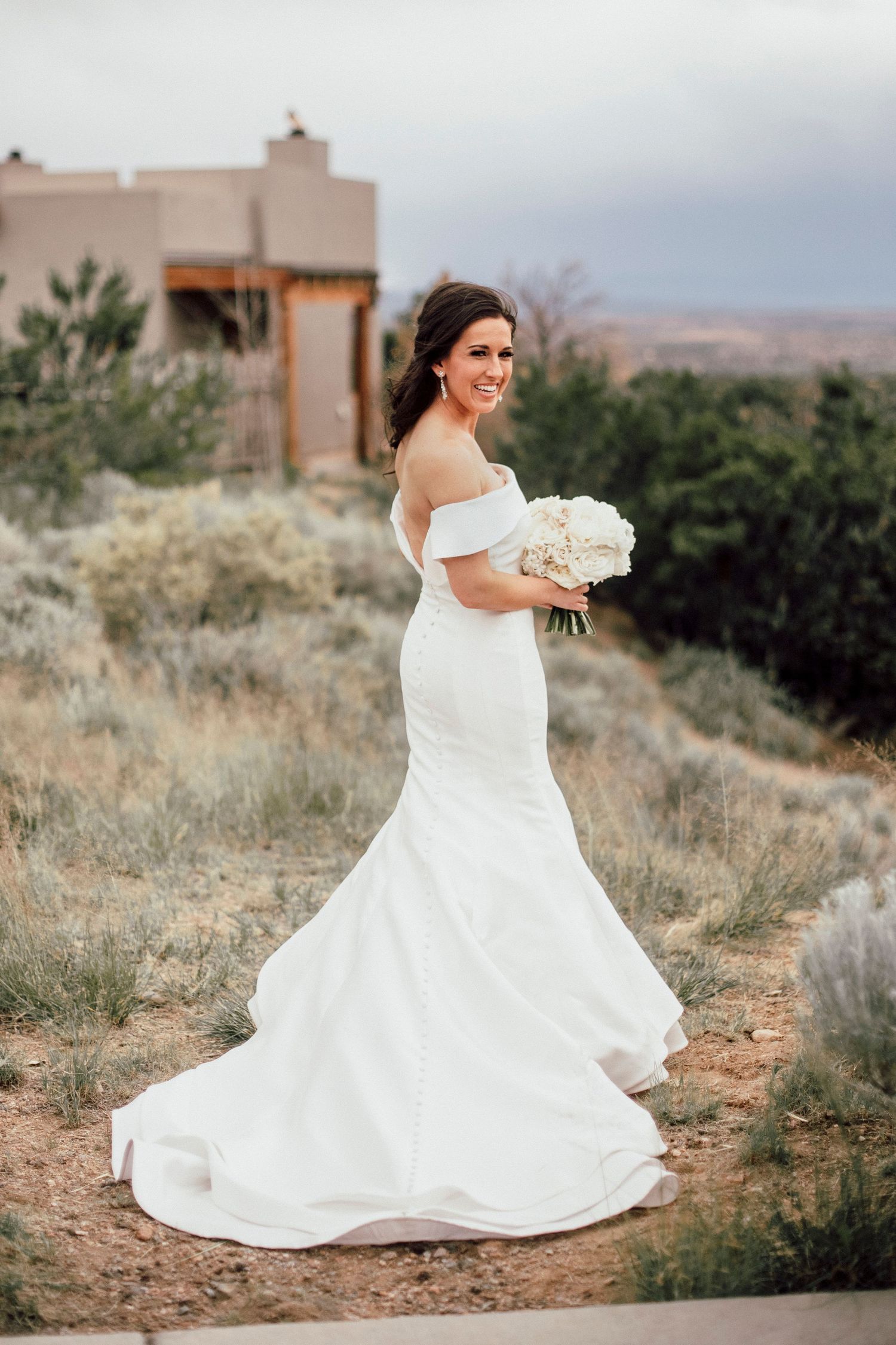 A bride in a fitted white mermaid wedding dress holds a bouquet while standing on a desert path with adobe buildings behind her.
