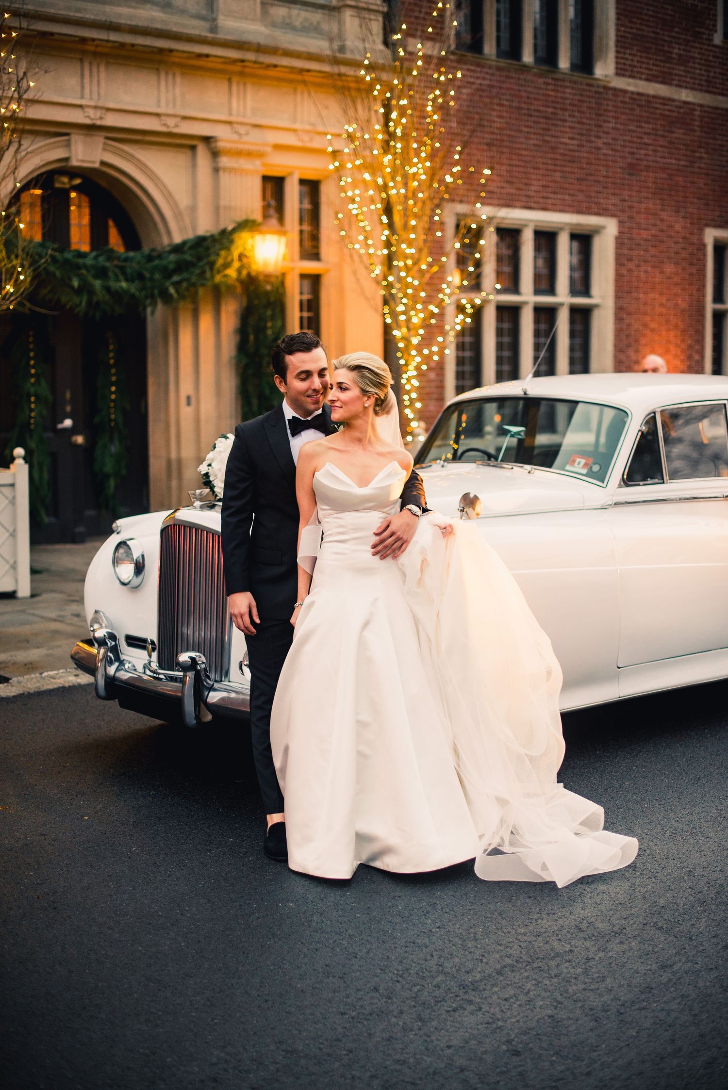 Newlywed couple shares romantic moments next to a classic white vintage car under twinkling lights at dusk.