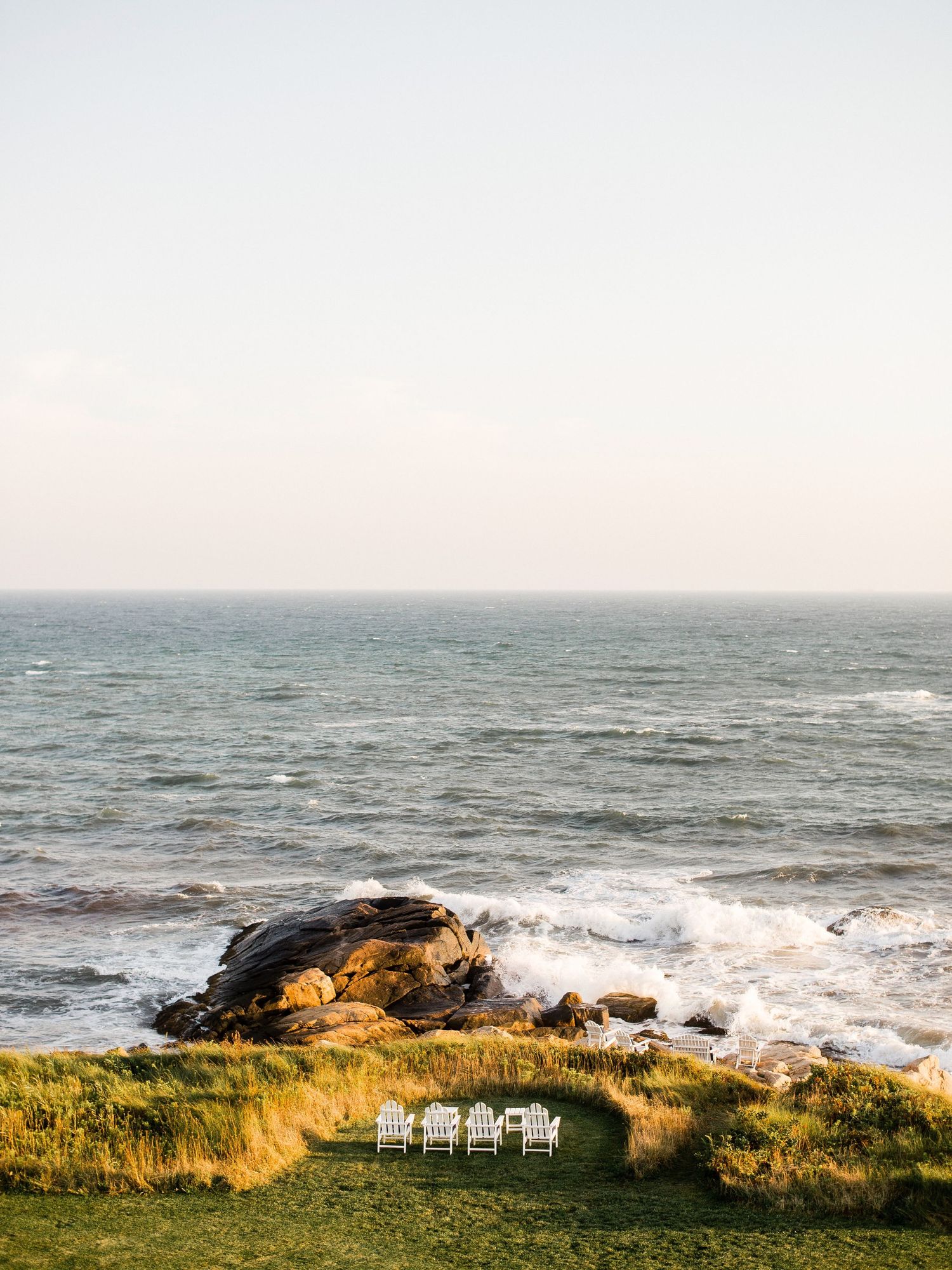 Coastal landscape in Little Compton RI with white chairs overlooking rocky shore and crashing waves at golden hour.