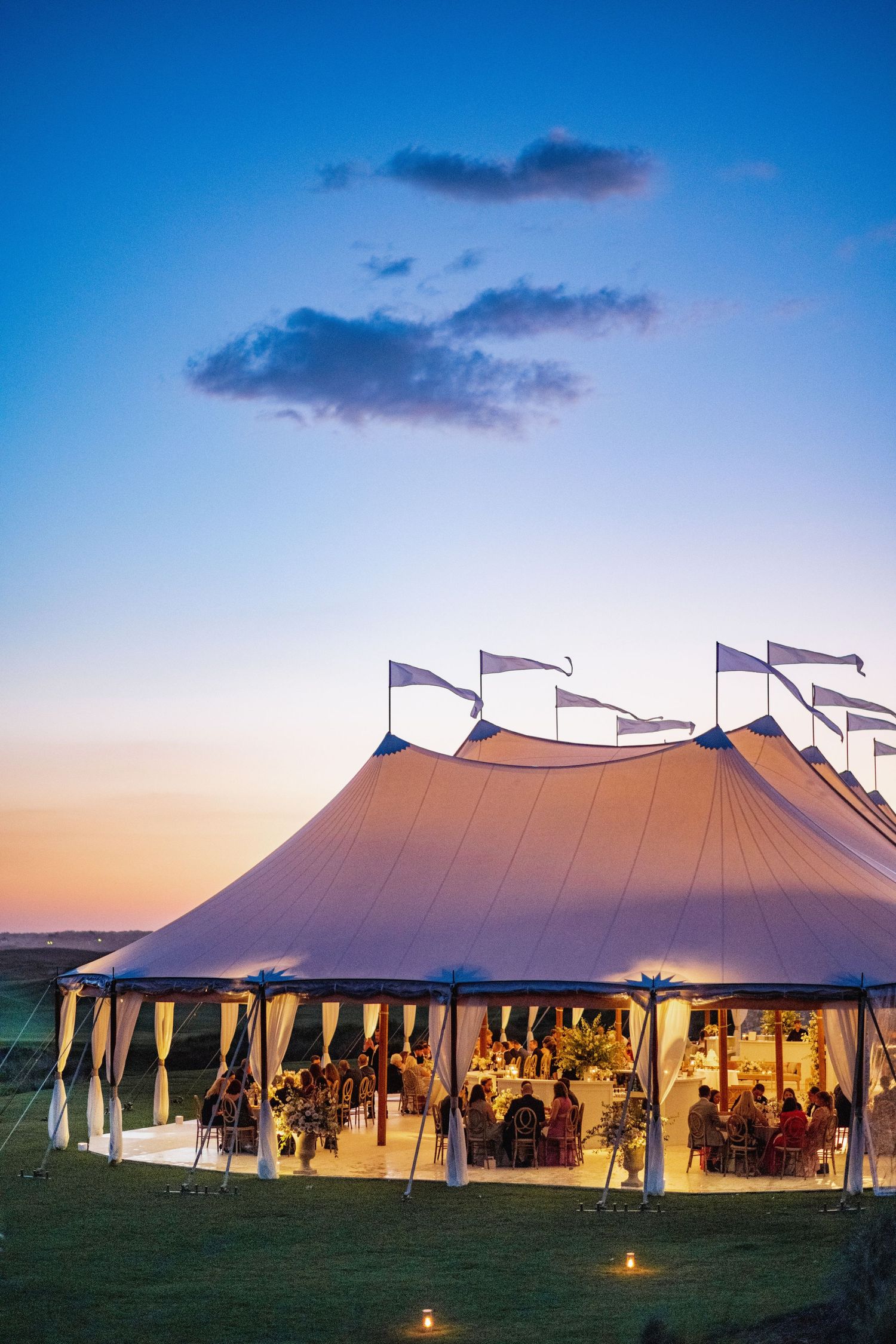 Large white wedding tents illuminated at dusk with flags flying on top at the Ocean Course, Kiawah