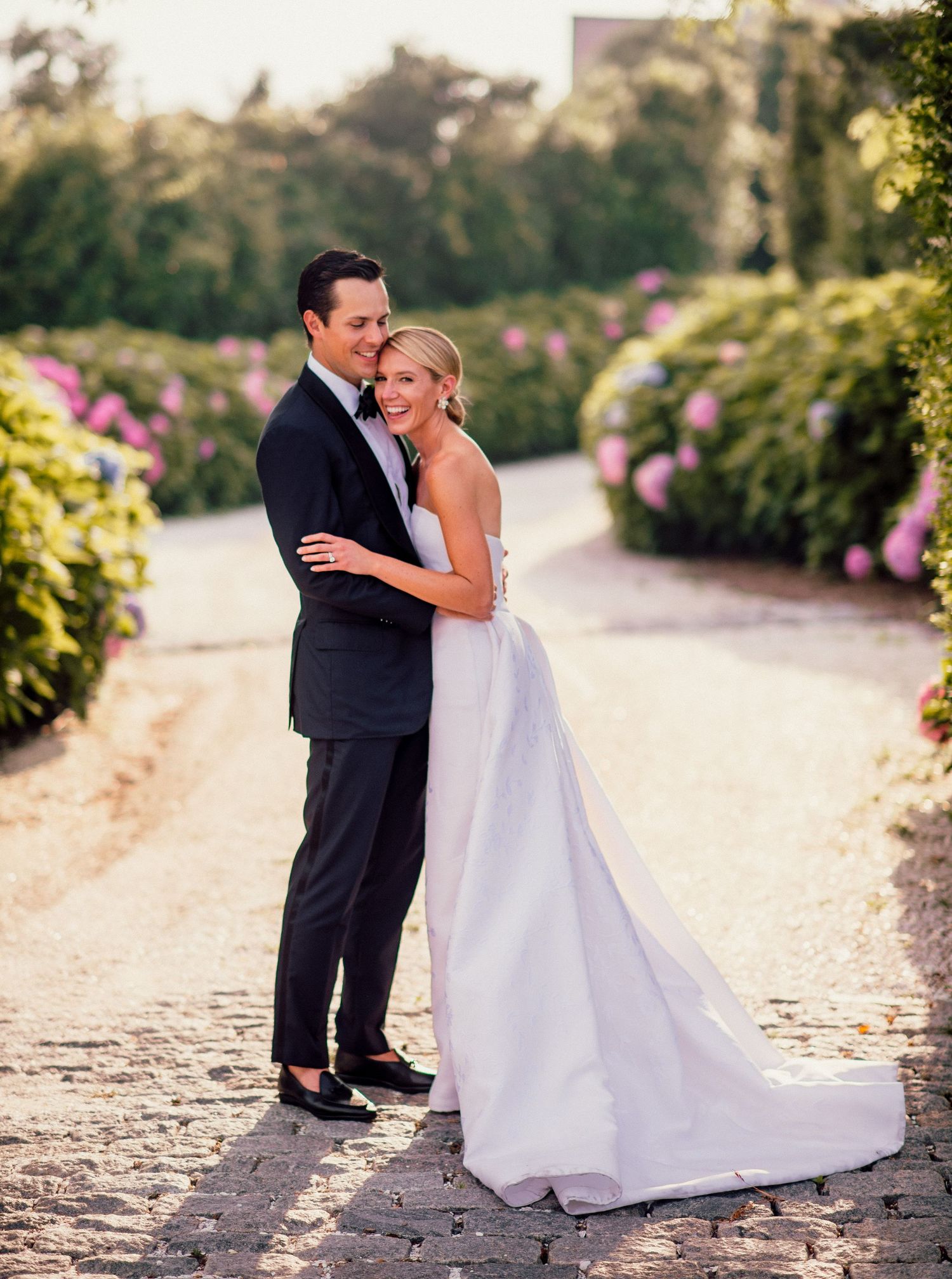Couple in wedding attire pose together on a garden path lined with pink flowers at sunset.