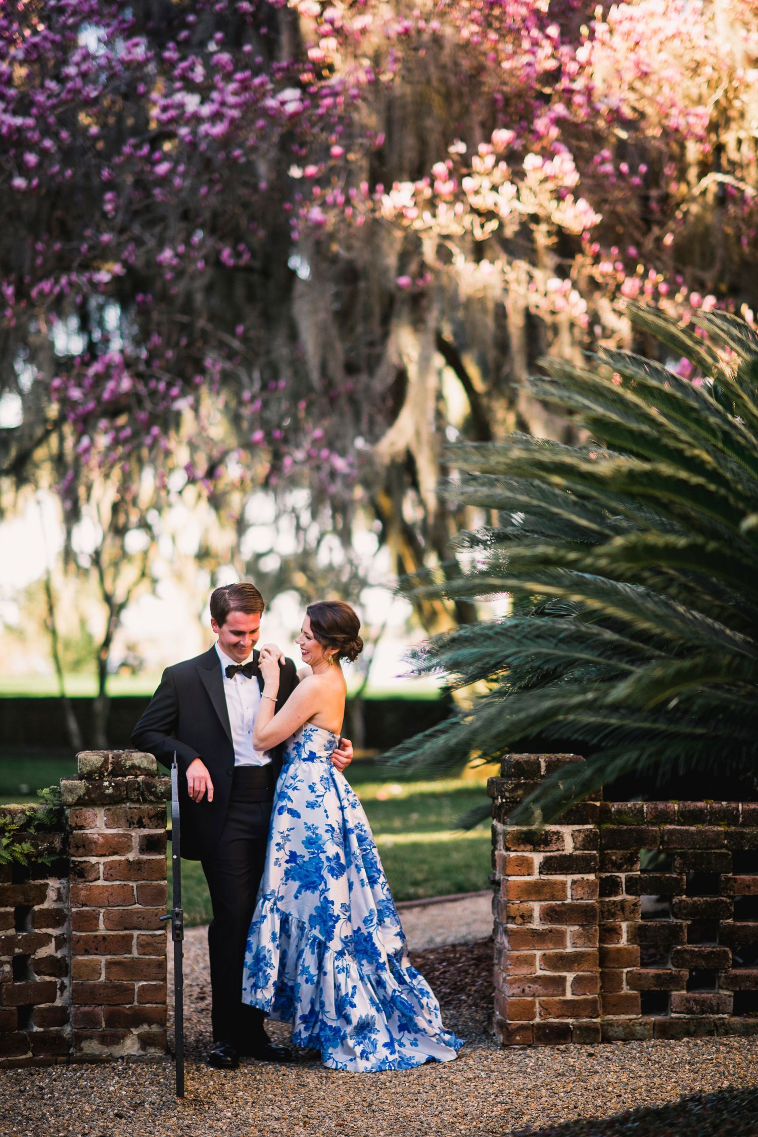 Couple in formal attire sitting by brick wall under flowering trees and Spanish moss.