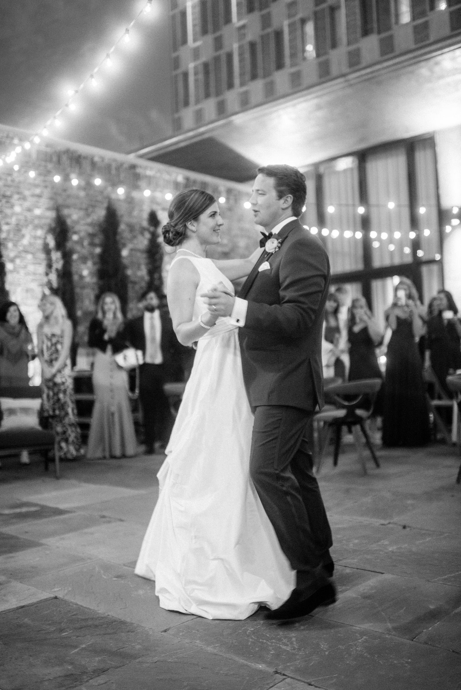 A romantic first dance sequence at an evening wedding reception under string lights captured in black and white photography.