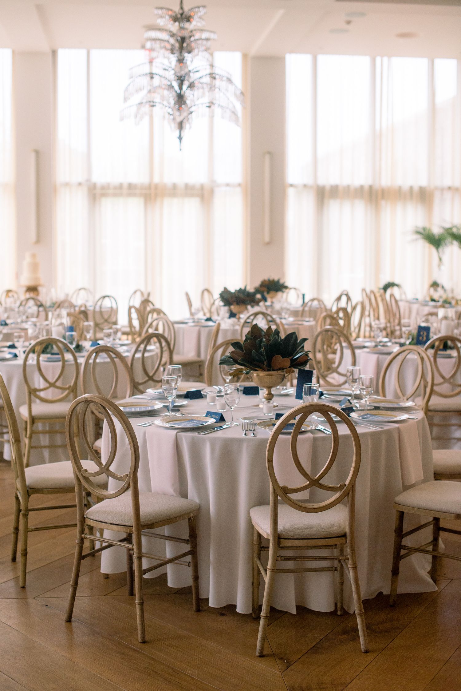 Elegant wedding reception venue with crystal chandeliers, white tablecloths, and gold infinity chairs in a bright room.