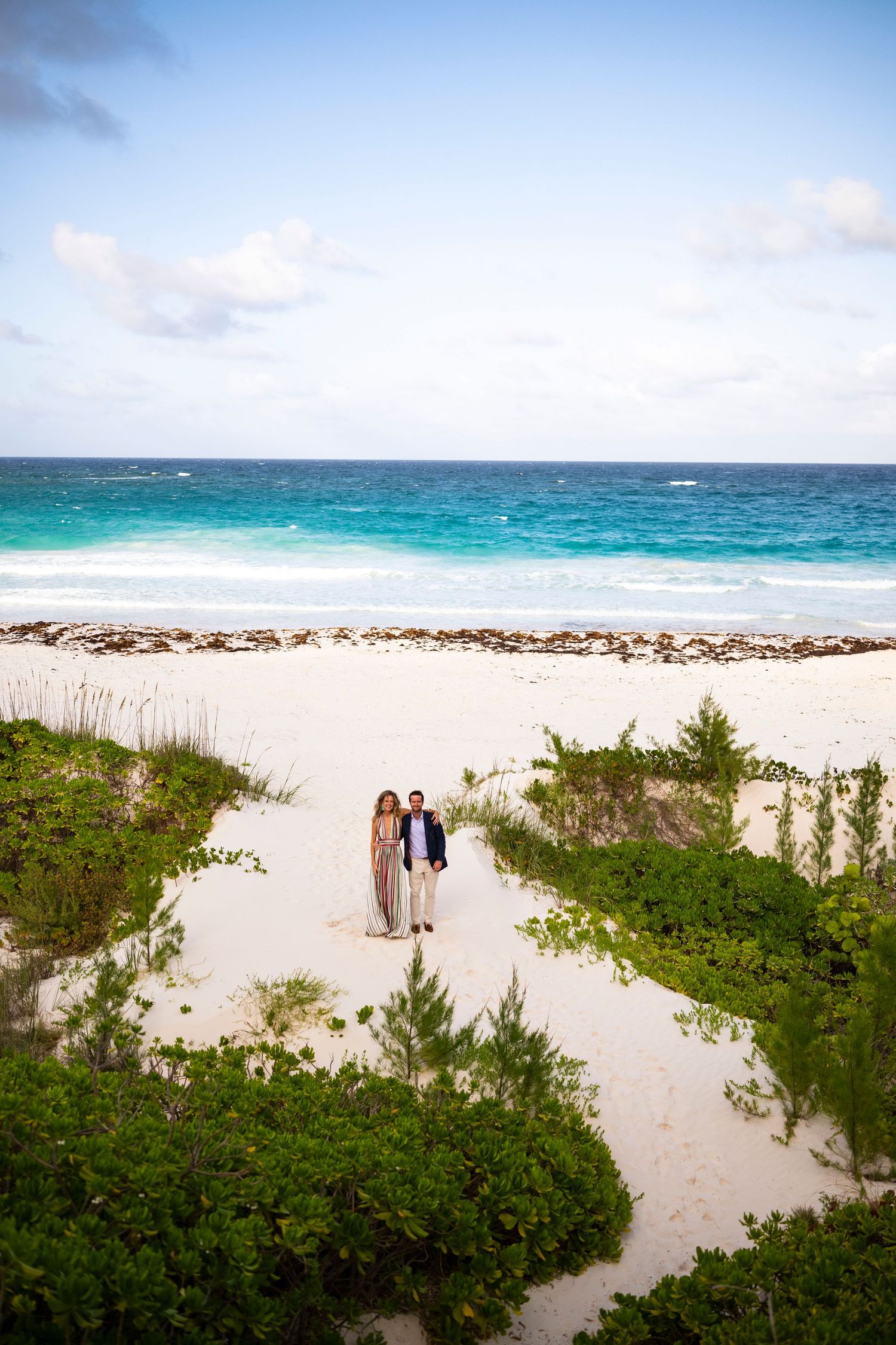A couple stands on a sandy path between green foliage with turquoise ocean waters and blue sky in the background.