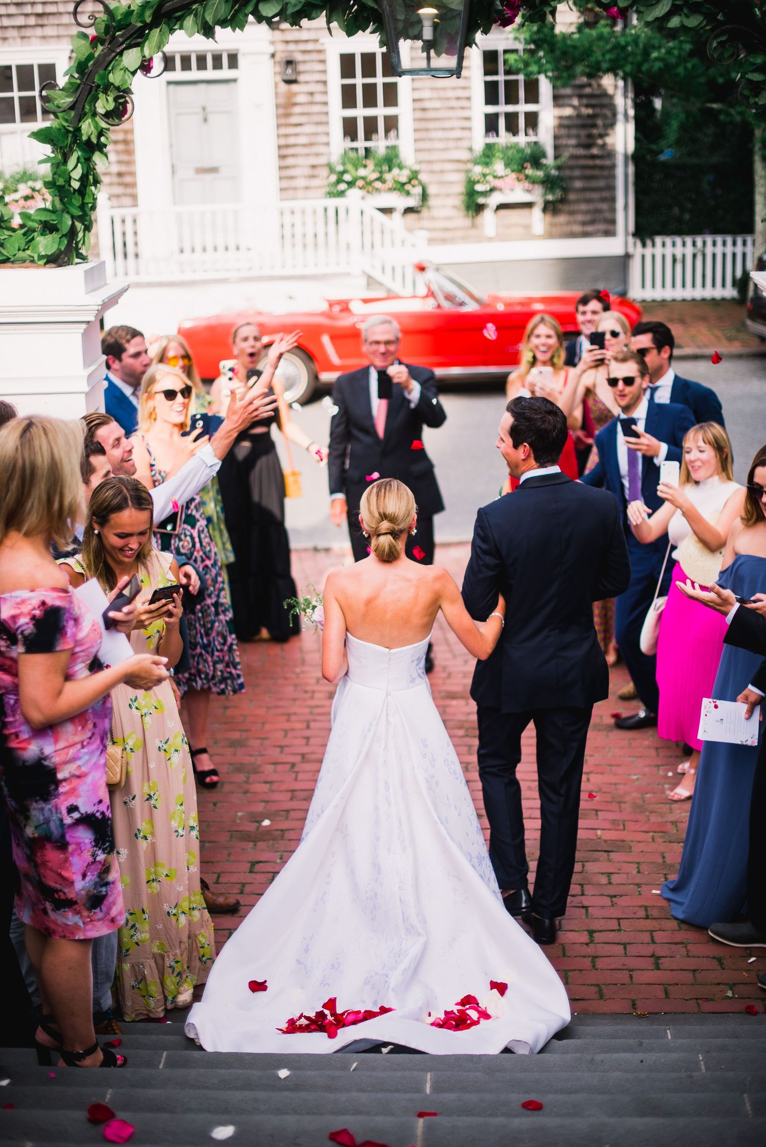 Wedding celebration on brick pathway with guests throwing petals and classic red car in background.