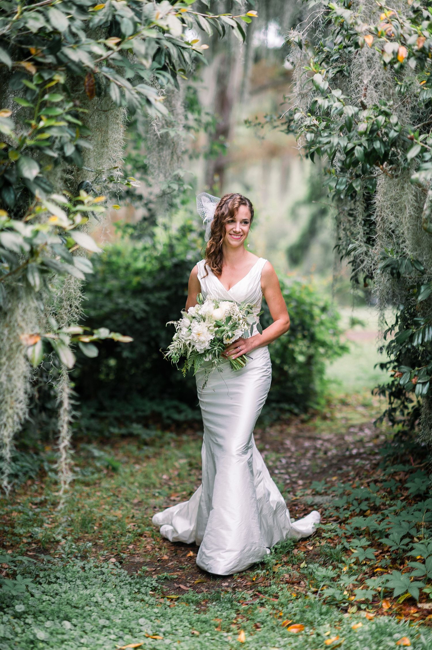 A white wedding dress stands out against lush greenery and Spanish moss in a romantic garden setting.