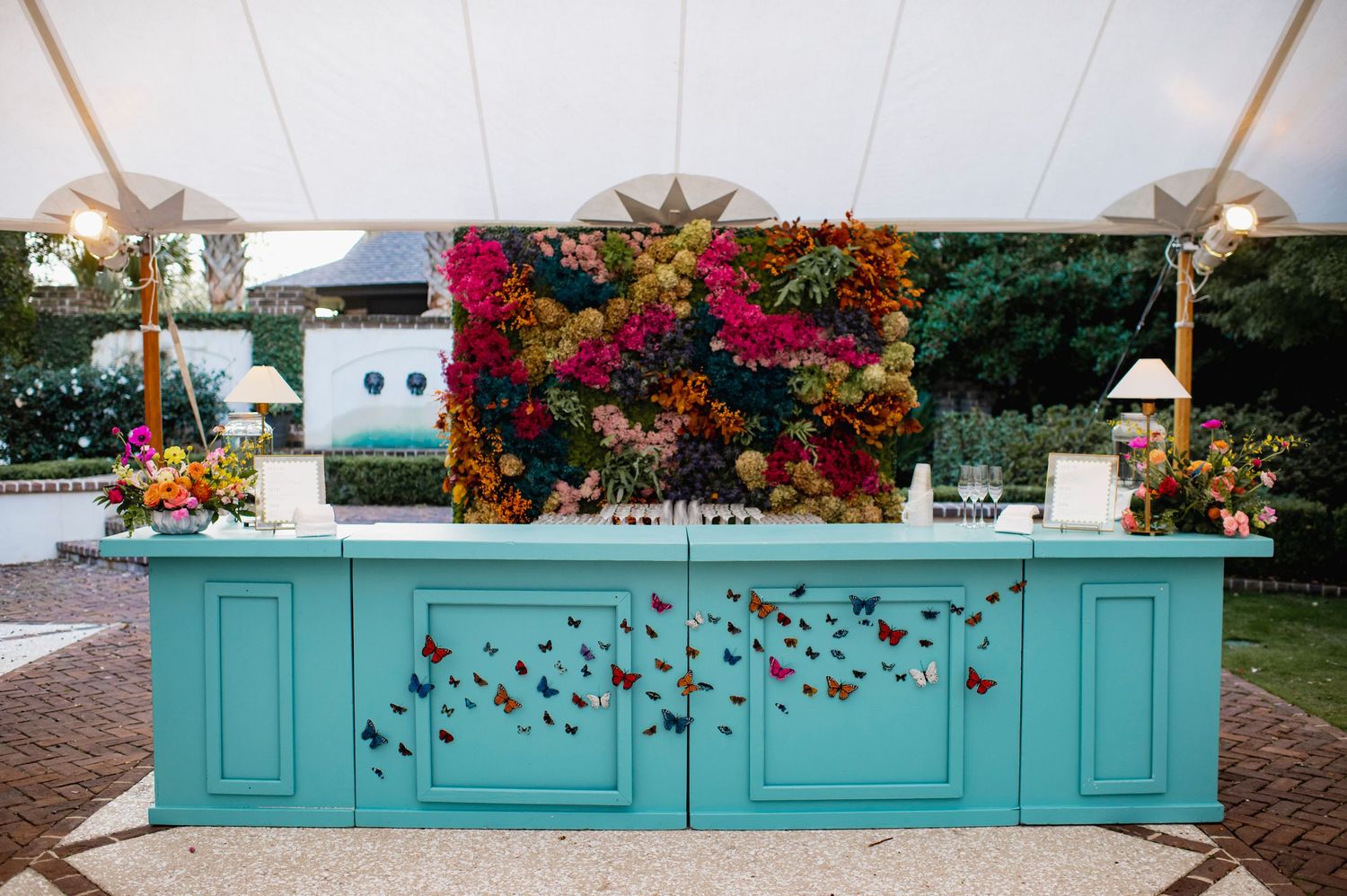 A turquoise bar decorated with butterflies sits in front of a vibrant floral wall display at an outdoor event.