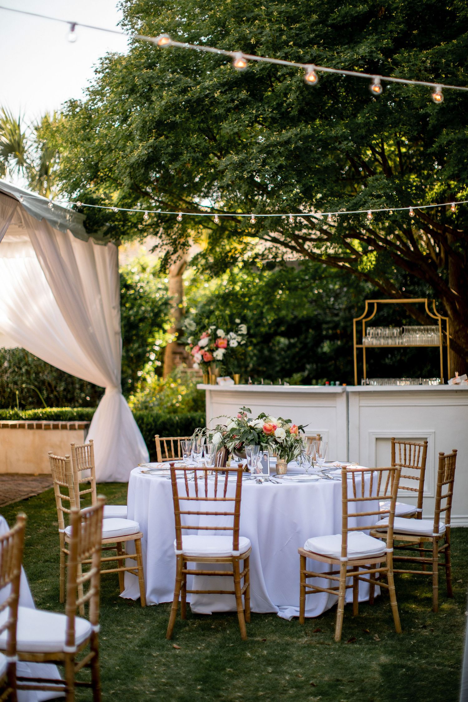 Elegant outdoor wedding reception setup with white linens, wooden chairs, and string lights hanging in garden setting.