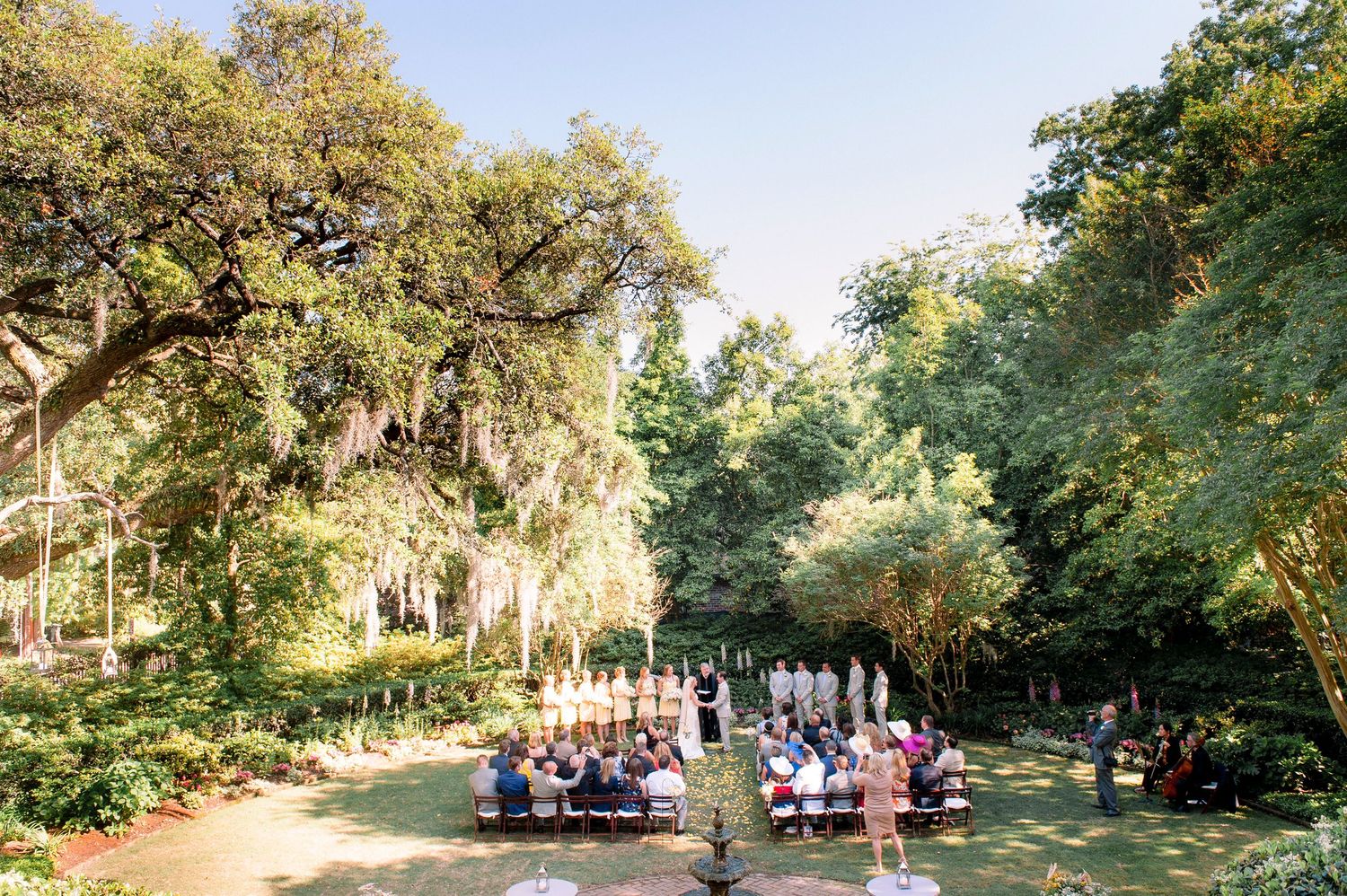 A panoramic view of an outdoor wedding ceremony setup with chairs arranged under large oak trees.