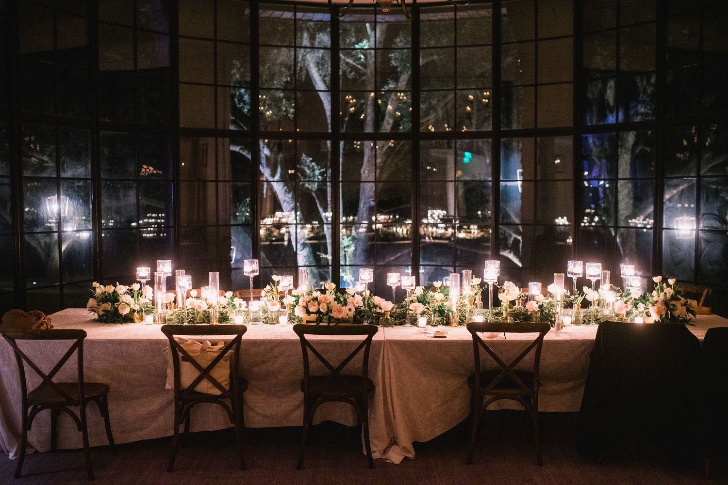 An elegant candlelit dining table set for a wedding reception with floor-to-ceiling windows overlooking city lights at night.