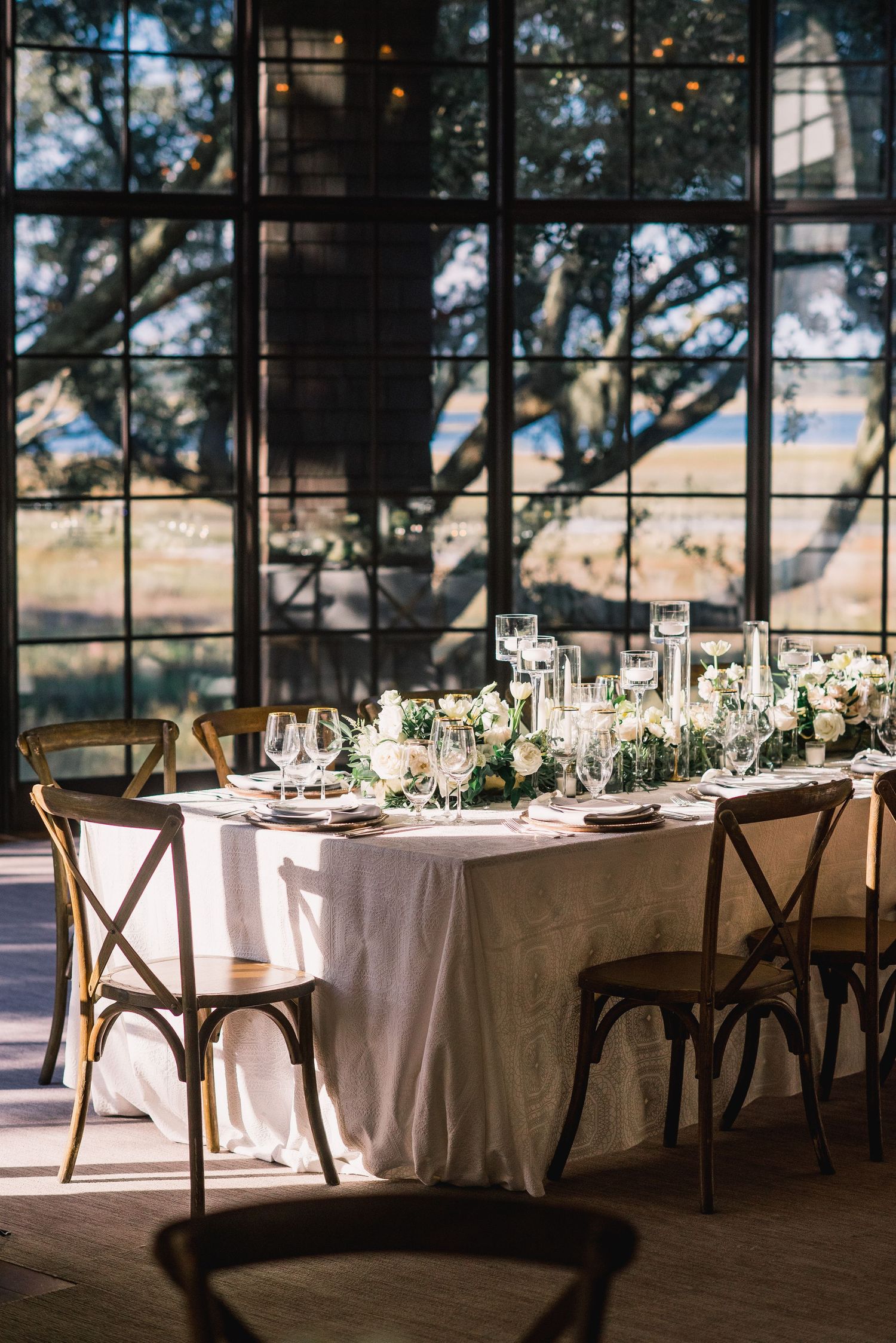 Elegant wedding reception tables with white linens and floral centerpieces set up in a sunlit venue with large windows.