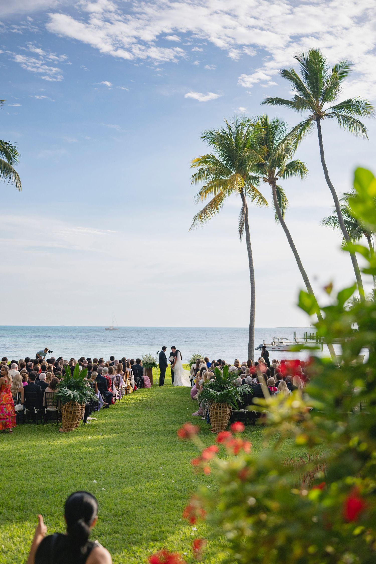 A beachside wedding ceremony setup with rows of white chairs on a green lawn overlooking the ocean and palm trees.