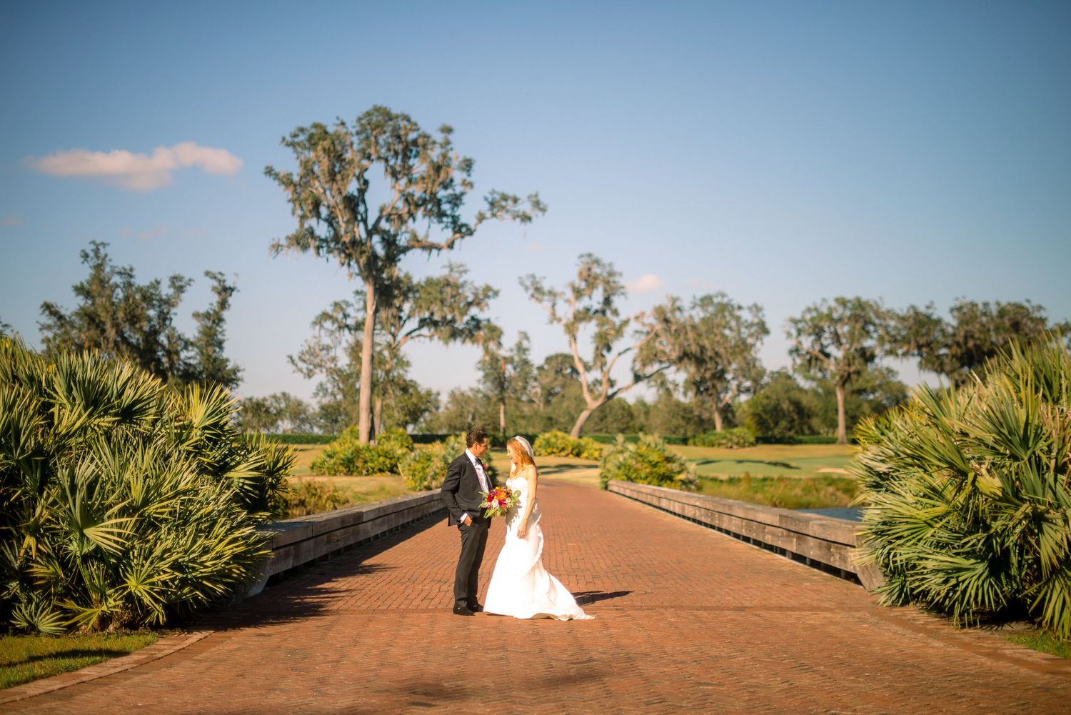 A couple in wedding attire poses along a scenic dirt road surrounded by lush tropical foliage and palm trees.
