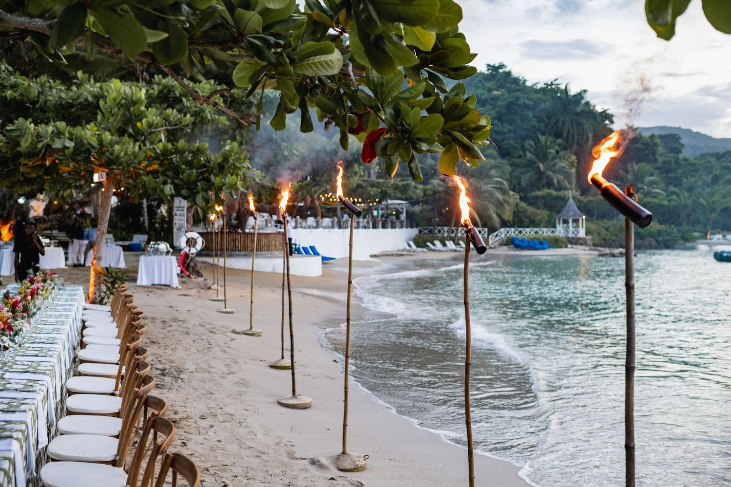 Flaming tiki torches line a beachfront walkway at sunset creating a tropical resort atmosphere by the ocean waves.
