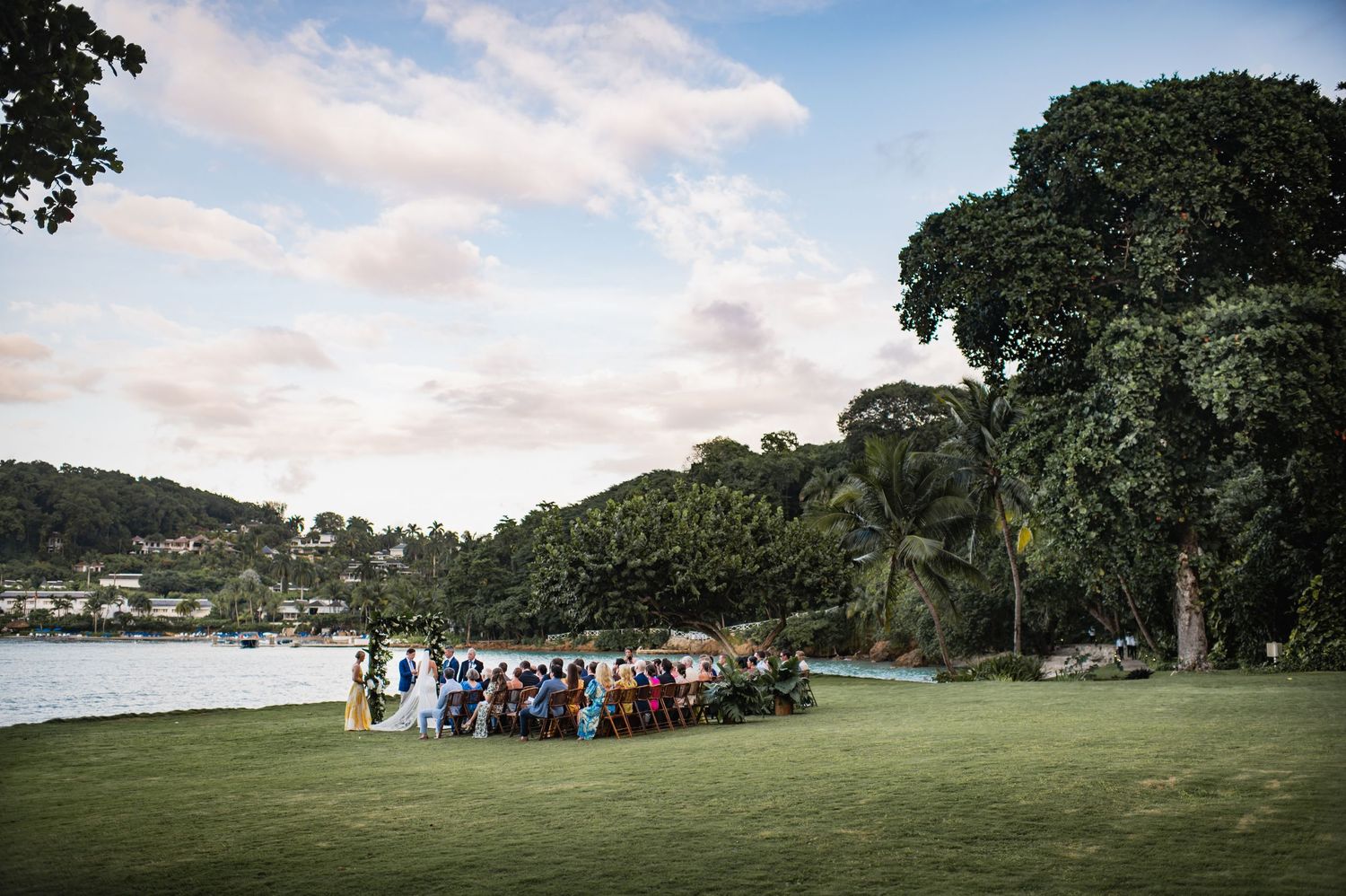A group of wedding guests gather on a lush green lawn overlooking a scenic bay with trees and cloudy skies at sunset.