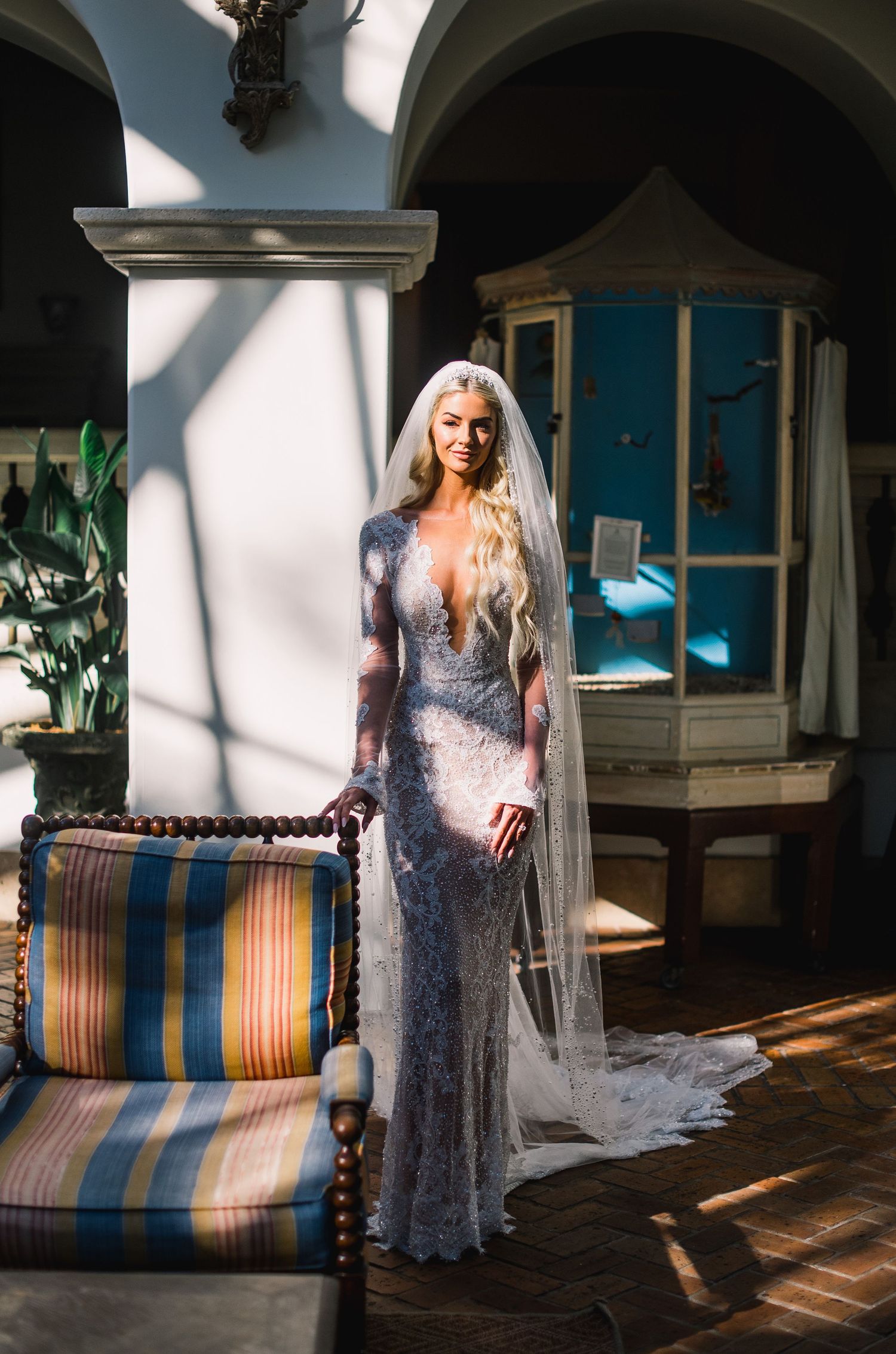 A bride in a sparkling purple gown stands in a sunlit room with striped vintage furniture and greenery.