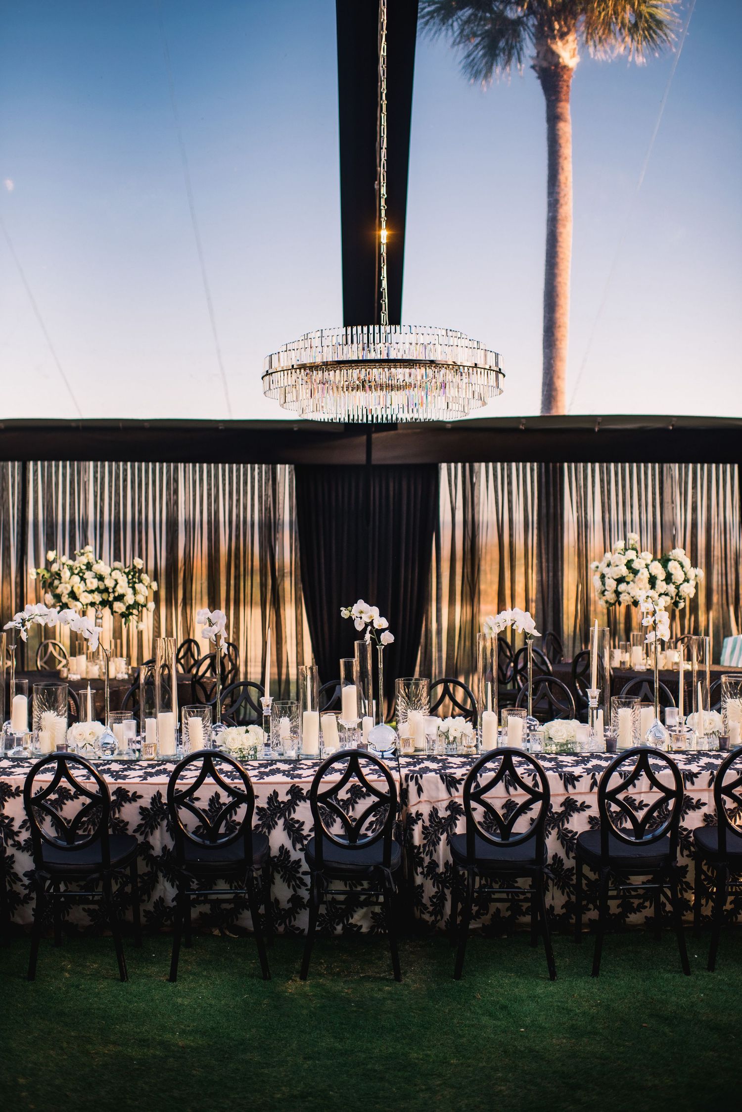 Elegant outdoor wedding reception setup with crystal chandelier, black drapes, ornate chairs and floral centerpieces at dusk.