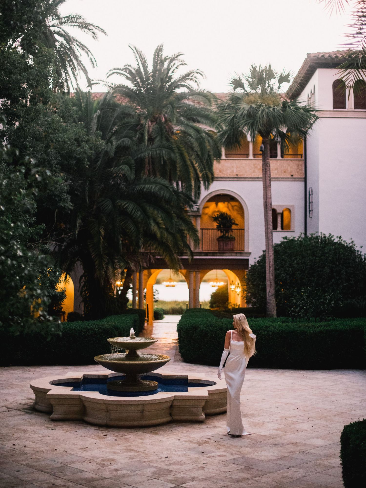 Panoramic view of a Mediterranean courtyard with palm trees and fountains at dusk.