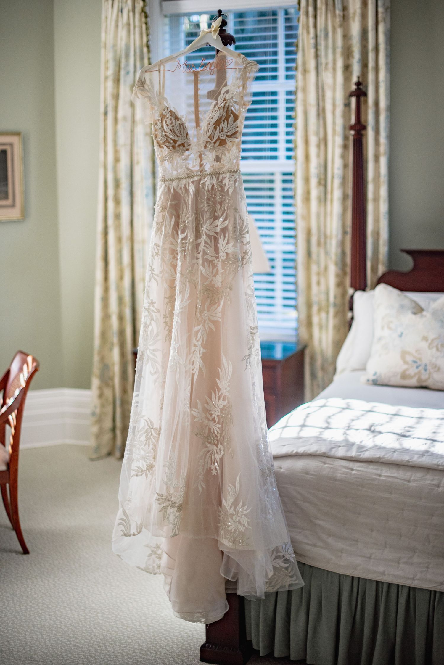 Delicate lace wedding dress hangs by window with white curtains in sunlit bedroom.