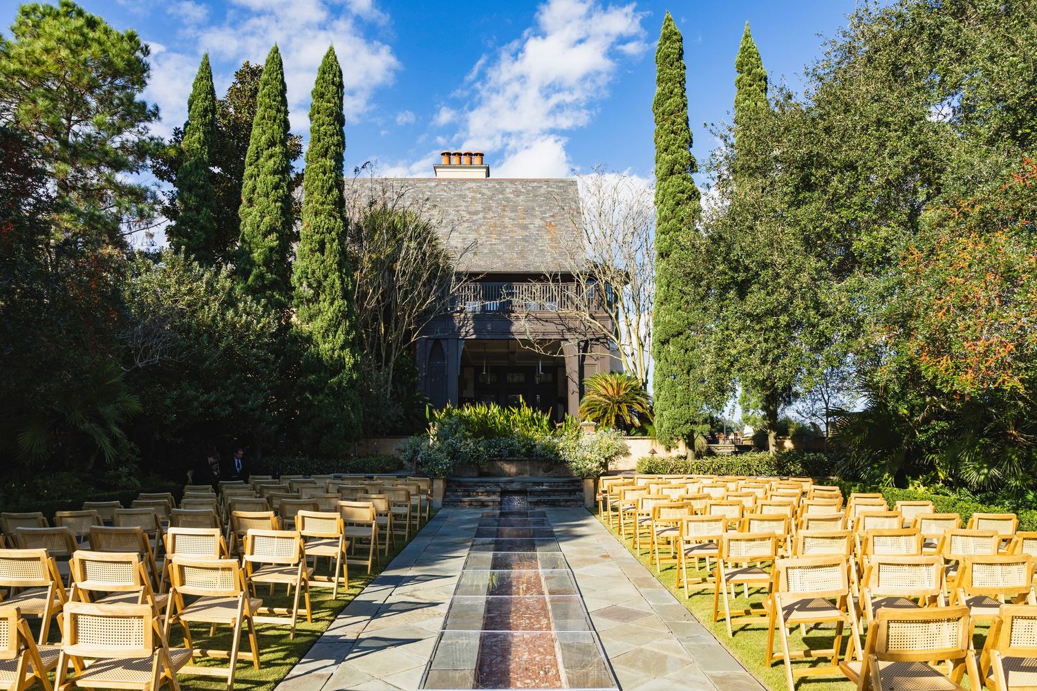 Outdoor wedding ceremony setup with wooden chairs arranged in rows along a stone path surrounded by lush gardens.