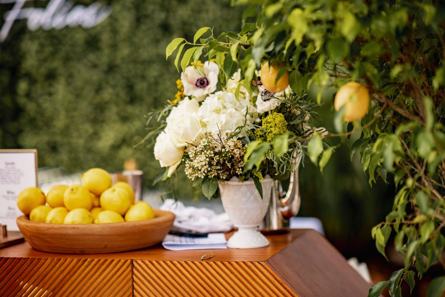 Fresh lemons and floral arrangements decorate an outdoor table setting with natural greenery in the background.