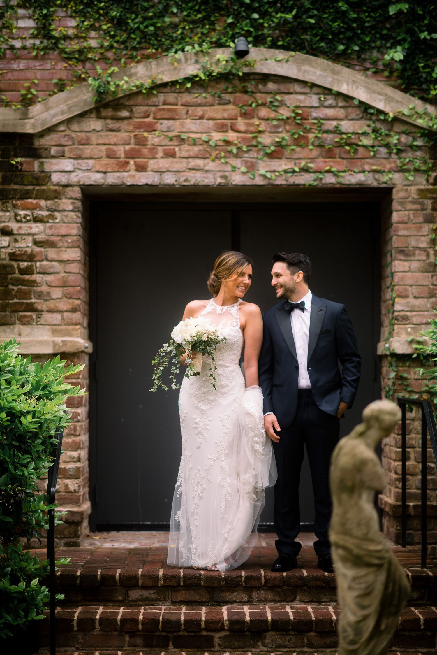 A romantic wedding moment captured in front of a brick archway with ivy, featuring elegant formal attire and a bridal bouquet.