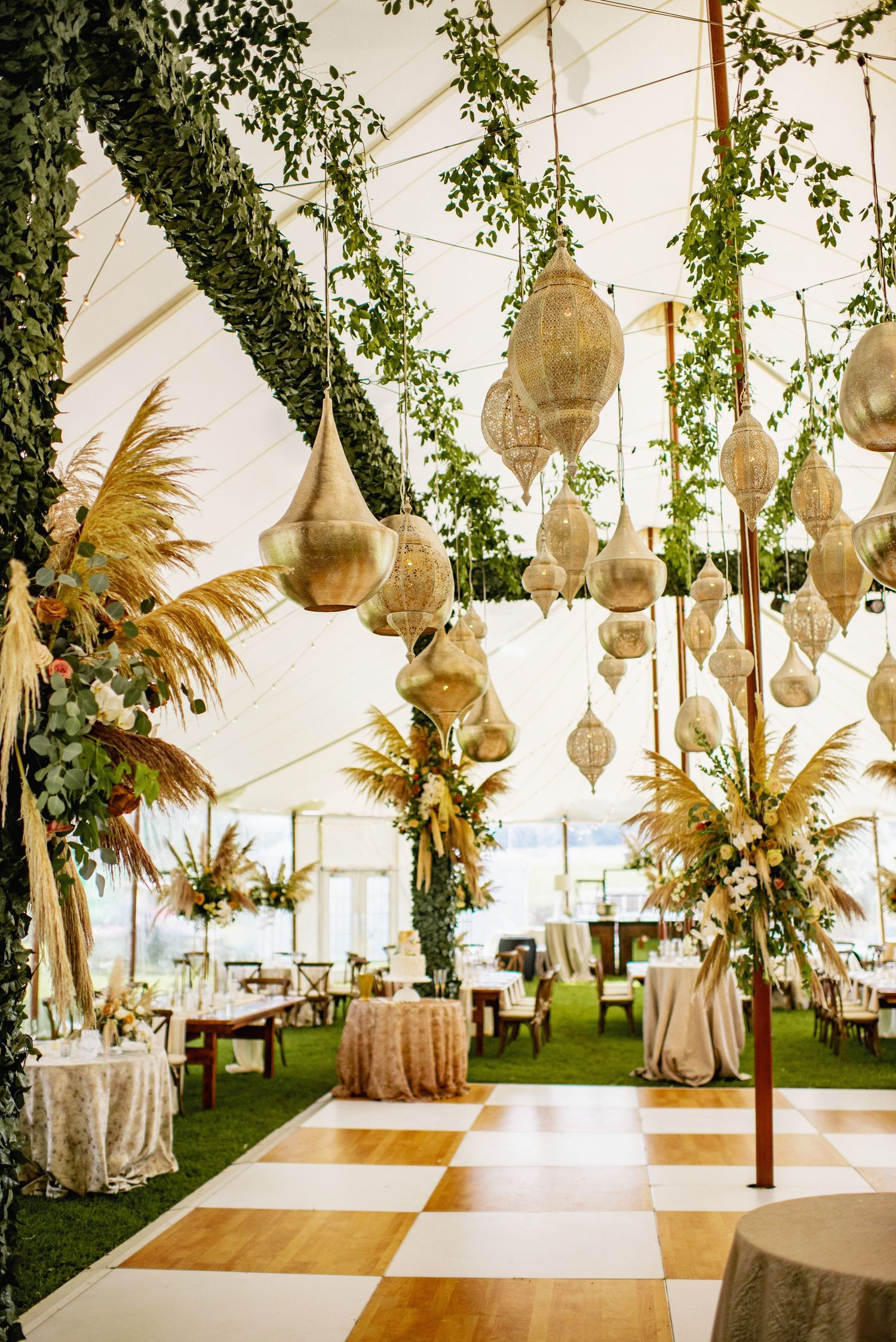 Elegant wedding reception tent with checkered dance floor, hanging gold lanterns and dried palm frond decorations.