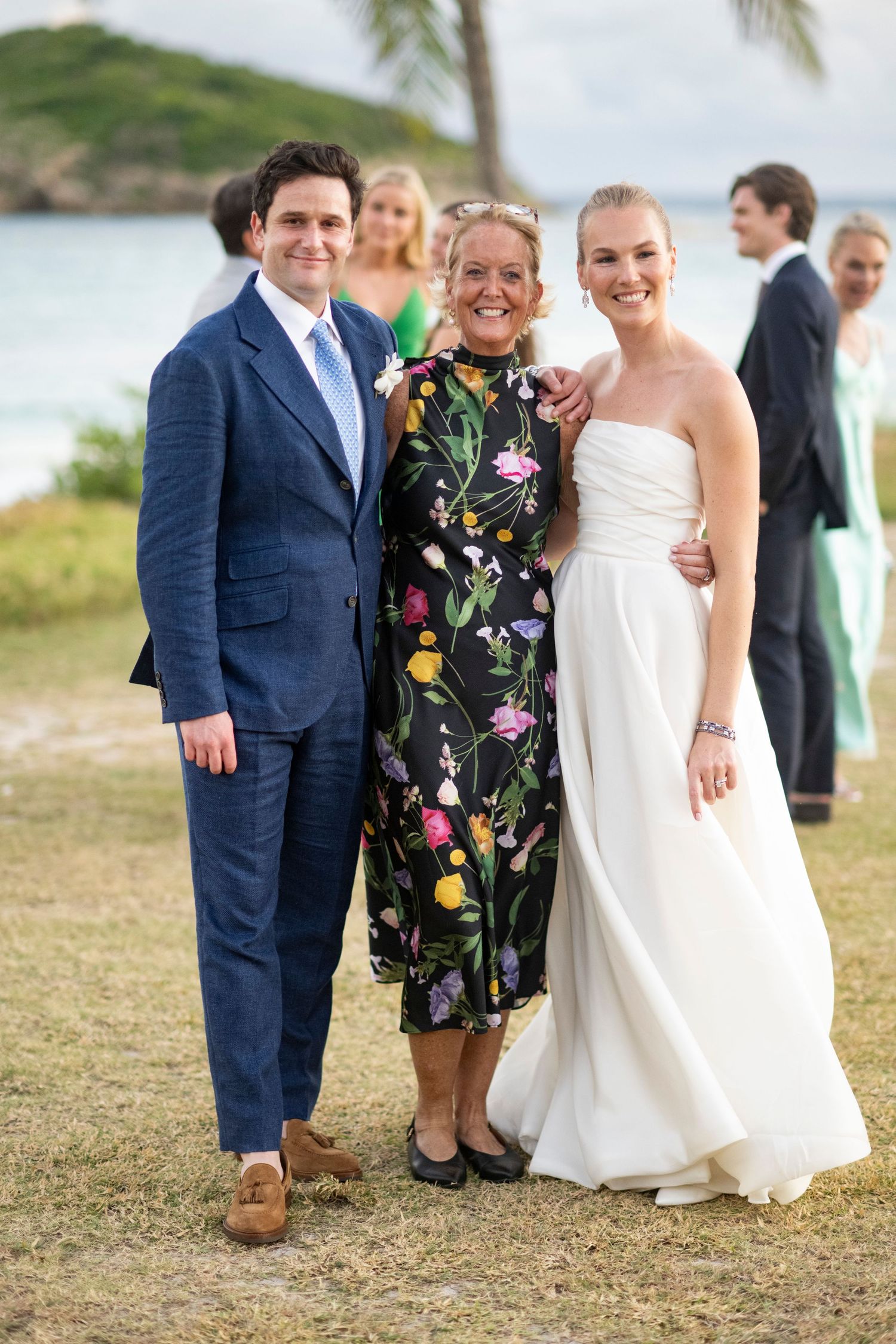 Wedding group photo on a grassy oceanside hill with palm trees in the background.