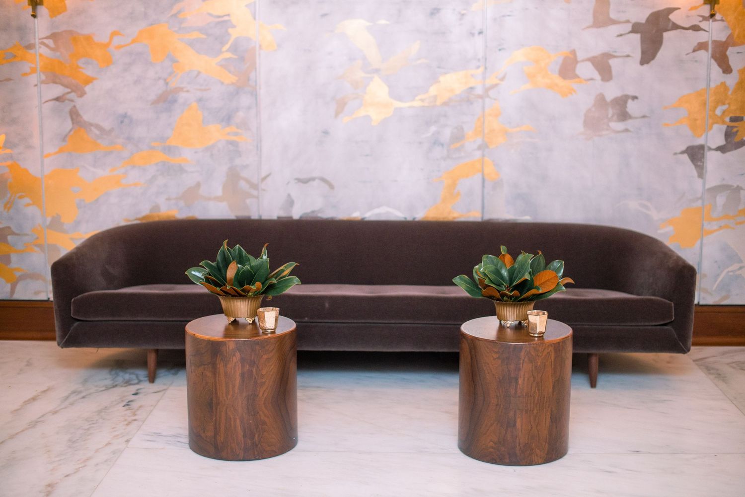 Modern living room decor with wooden accent tables, potted plants, and a gray sofa against a textured copper wall.