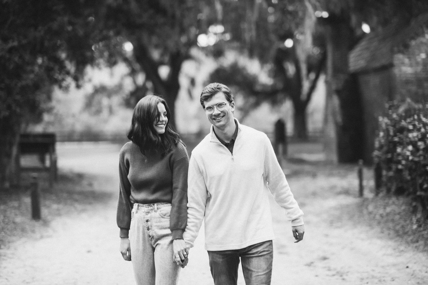 A couple walking together on a tree-lined path, holding hands and smiling in black and white.