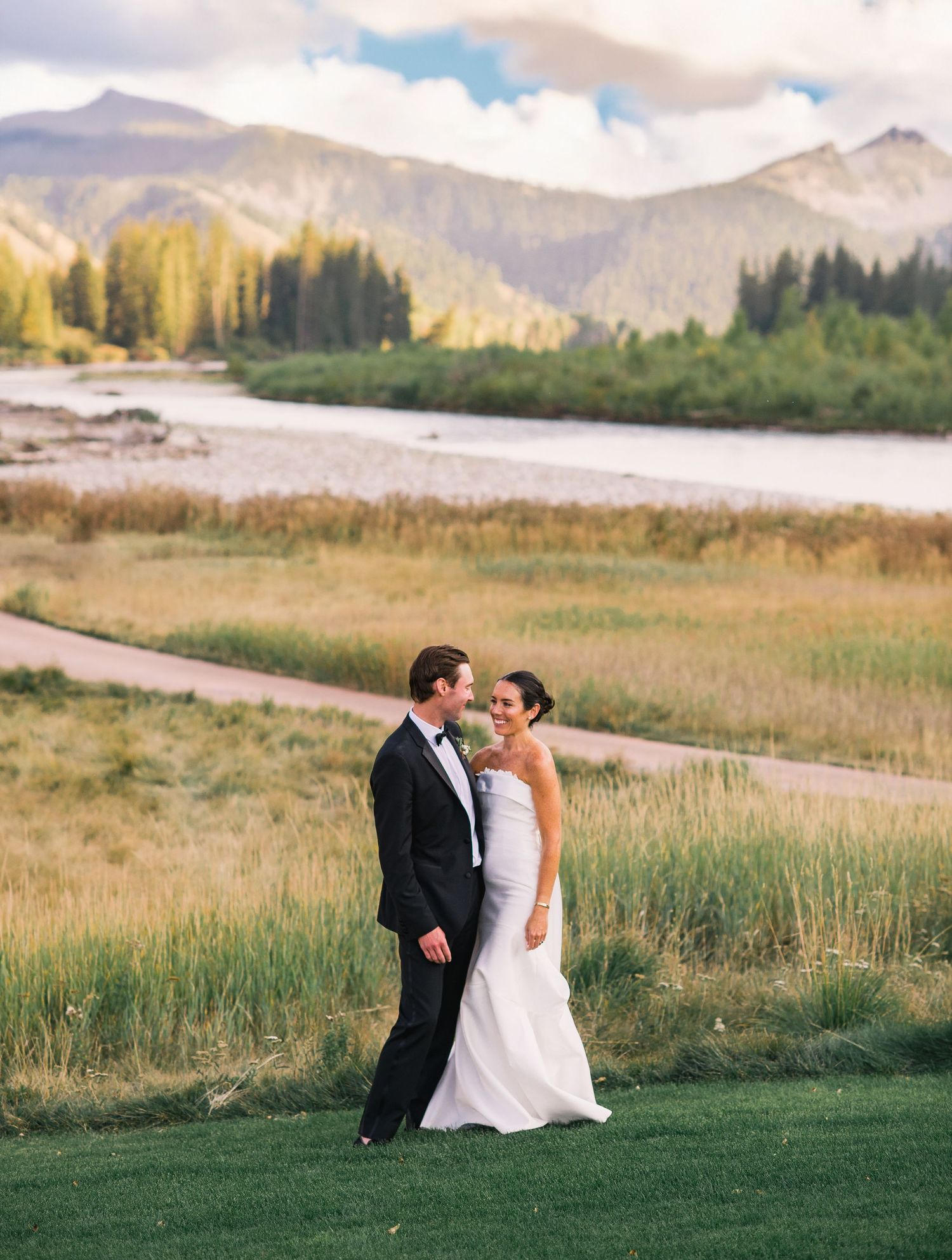 A couple in wedding attire stands together in a scenic mountain meadow with dramatic peaks in the background.