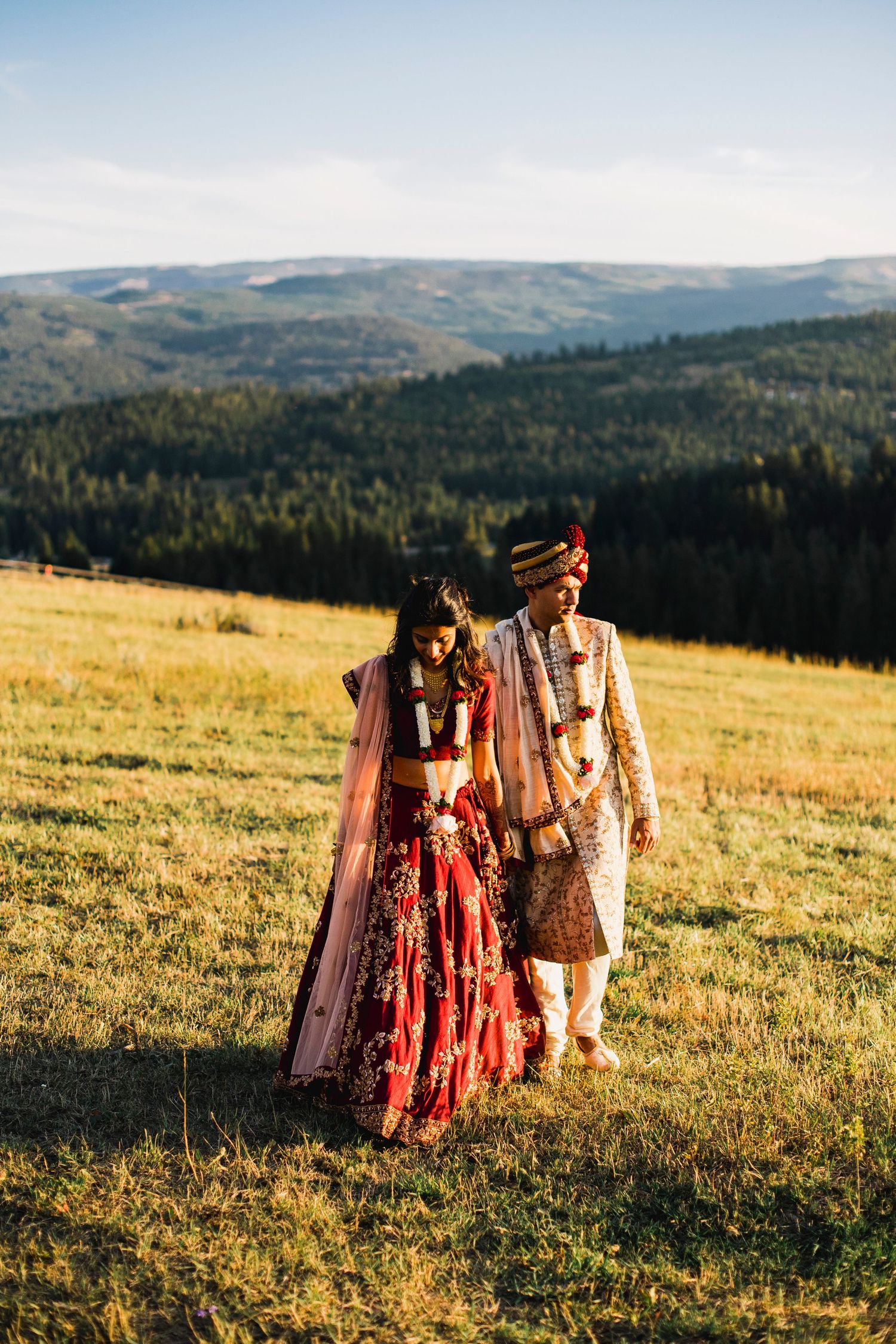 A couple in traditional cultural wedding attire walks through a sunlit mountain meadow at golden hour.