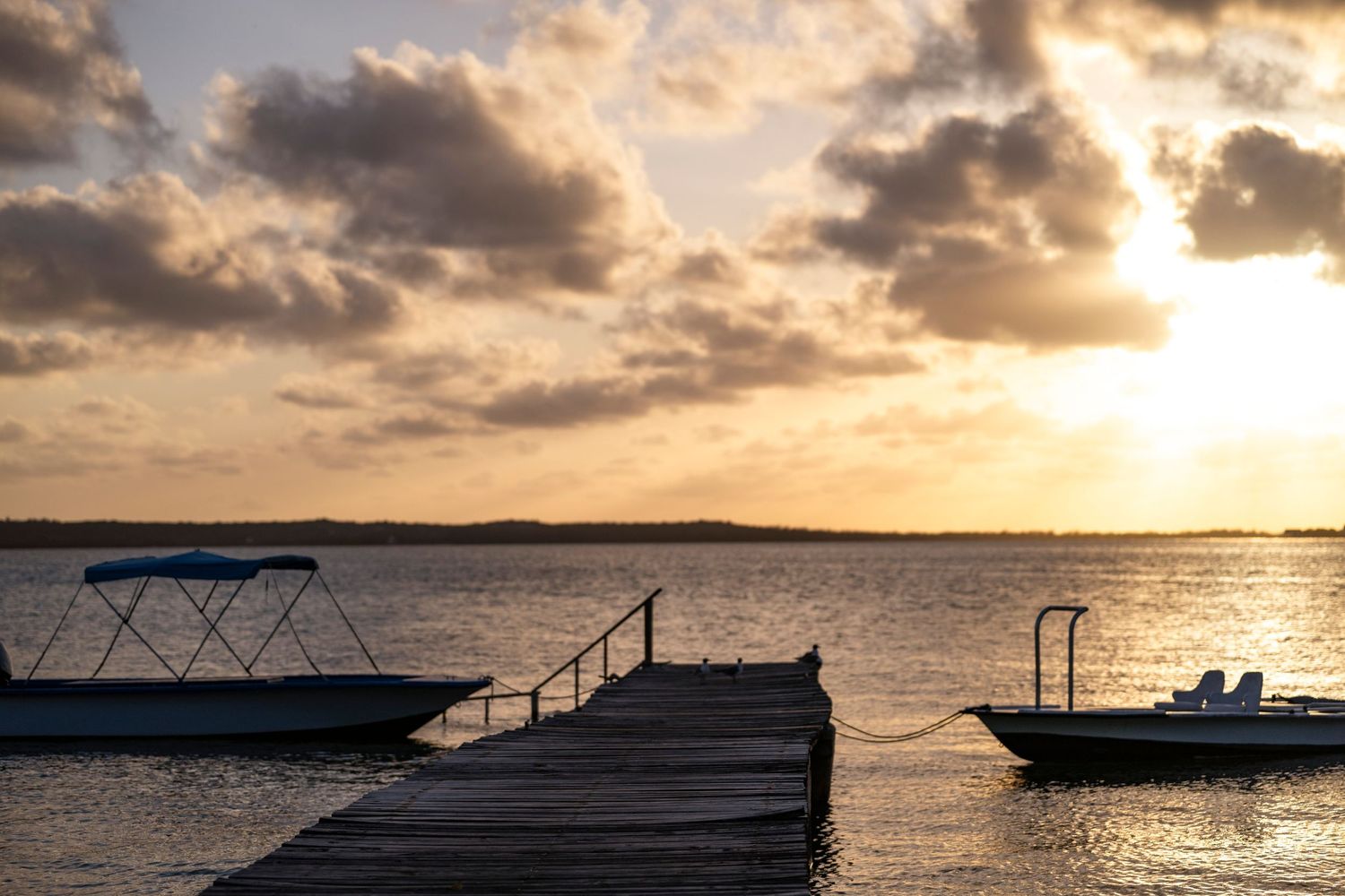 Wooden dock with boats silhouetted against dramatic orange sunset over calm waters.