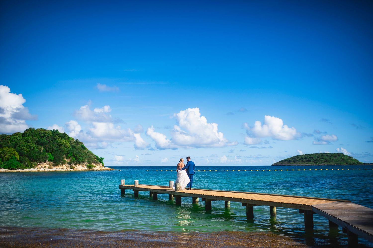 A wooden pier extends into turquoise waters under bright blue skies with tropical islands in the distance.