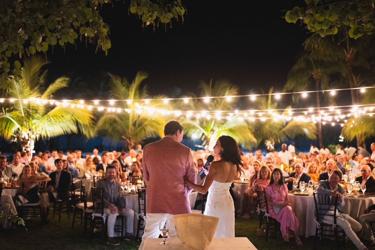 Outdoor evening event with string lights illuminating tables and palm trees creating a festive ambiance.