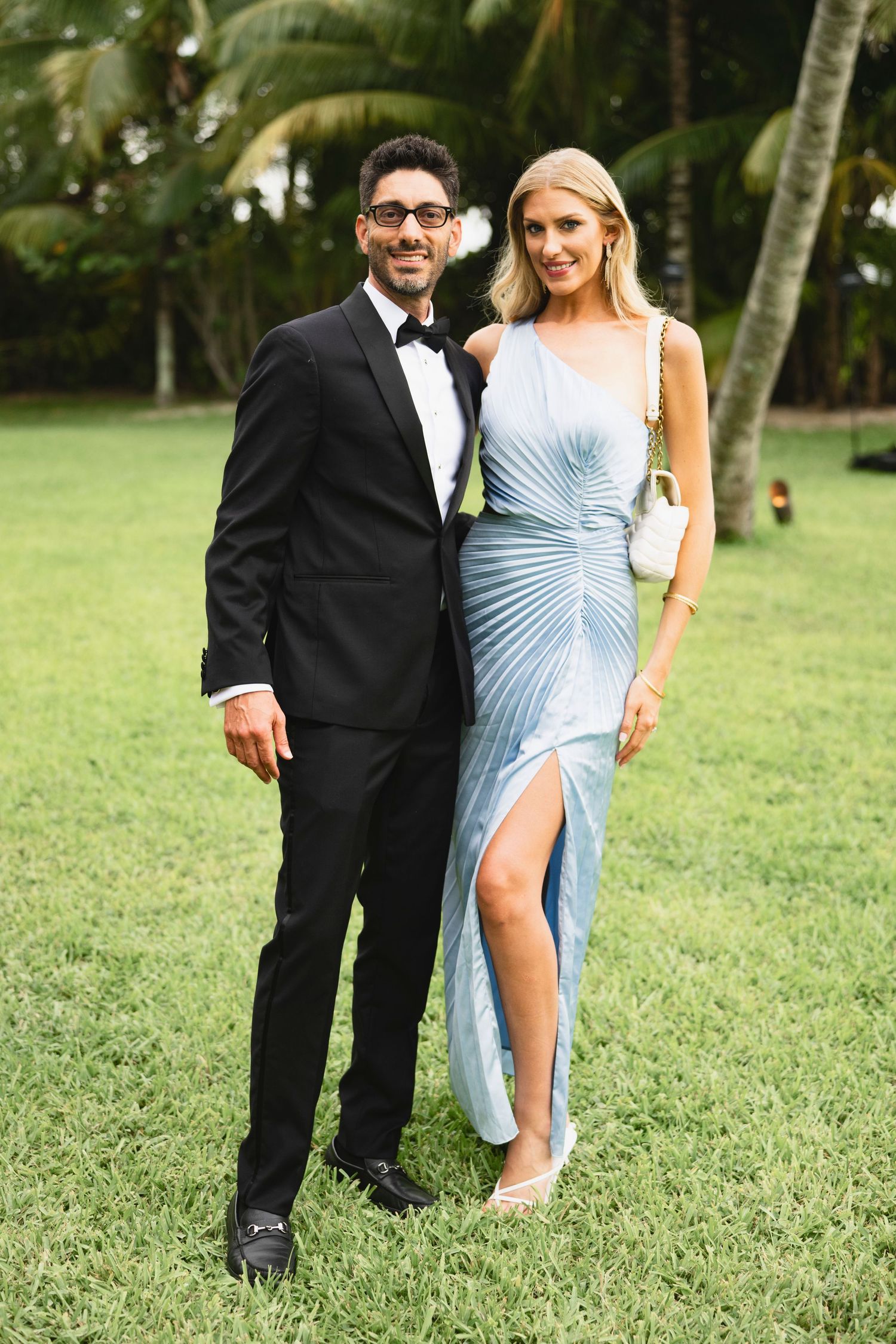 A couple poses on a lawn with palm trees, wearing formal attire - him in a tuxedo and her in a light blue gown.