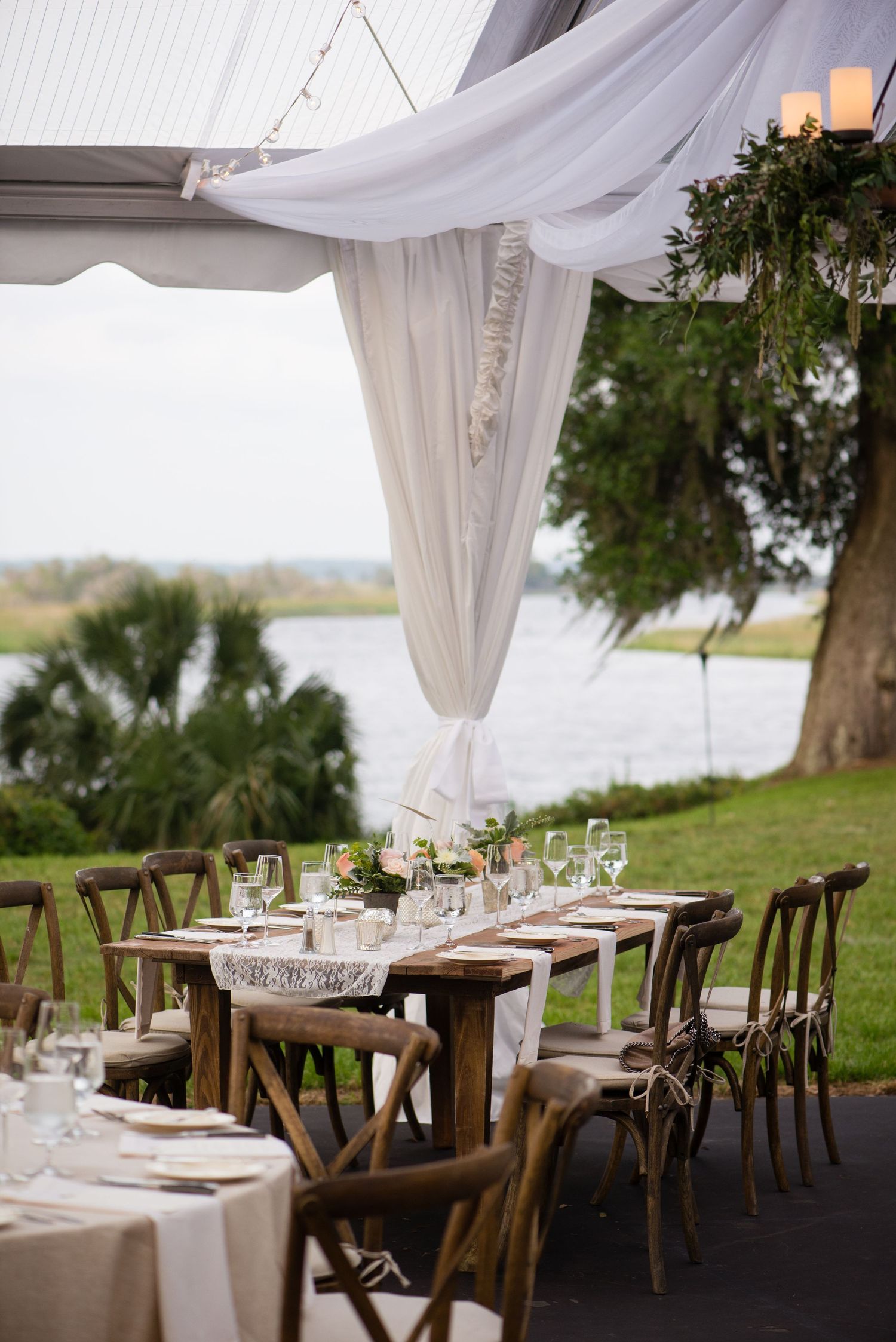 Elegant outdoor wedding reception setup with white drapes, wooden chairs and tables overlooking a scenic lake.