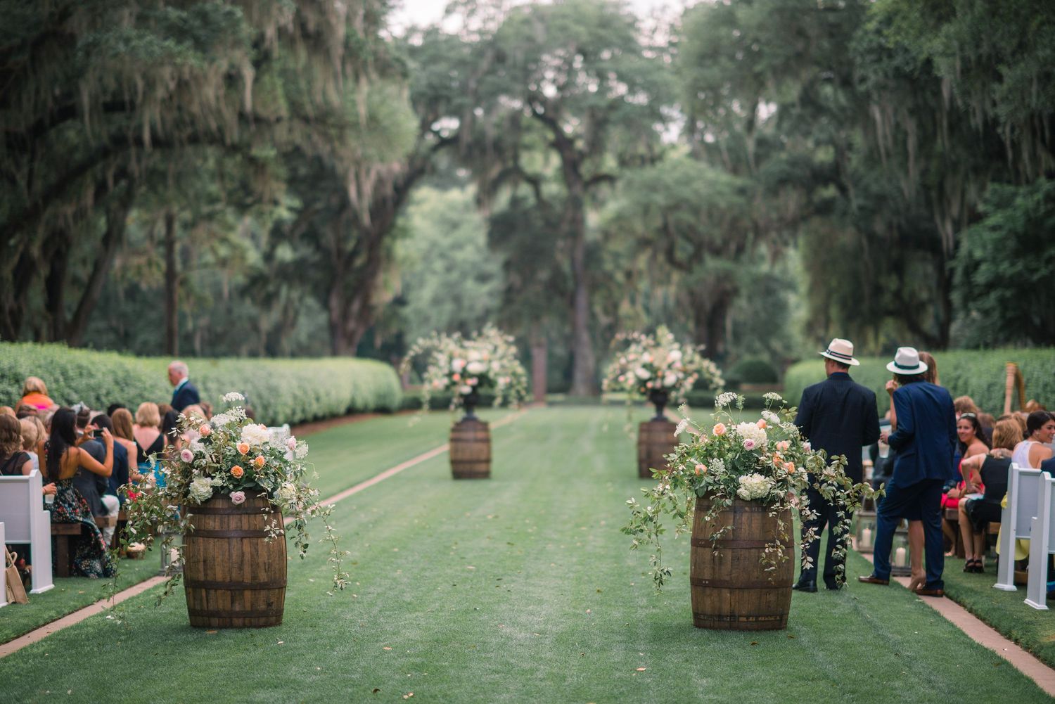 Rustic wooden barrels line a lush green outdoor wedding aisle decorated with Spanish moss and trees.